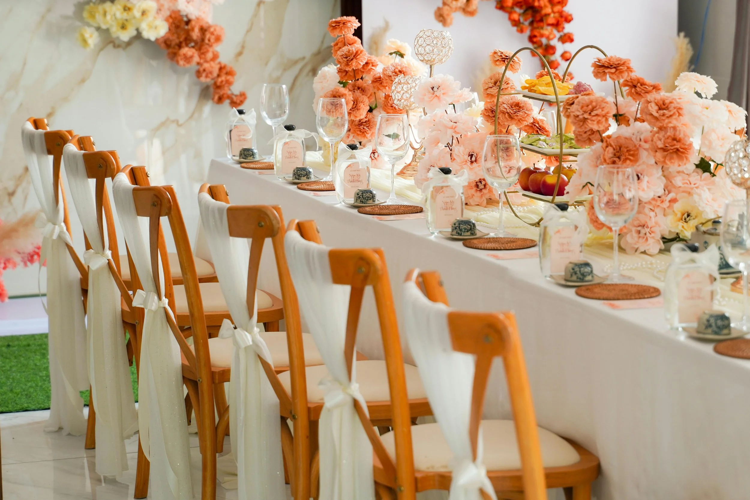 A beautifully decorated banquet table with pink and peach floral centerpieces, wine glasses, and place settings with small tags and place mats. The chairs are wooden with white fabric sashes tied on the back.