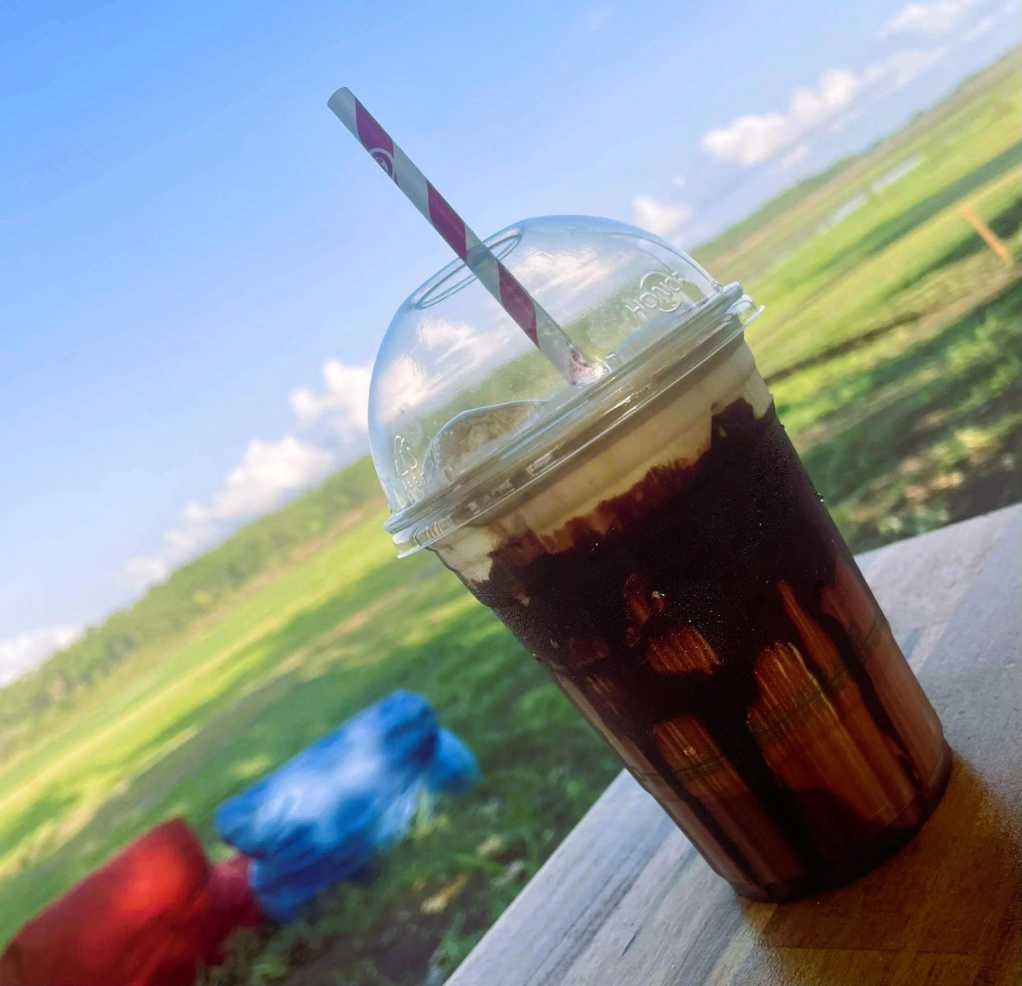 A cold coffee drink in a clear plastic cup with a dome lid and a striped straw, outdoors on a wooden surface with a scenic green field and blue sky in the background.