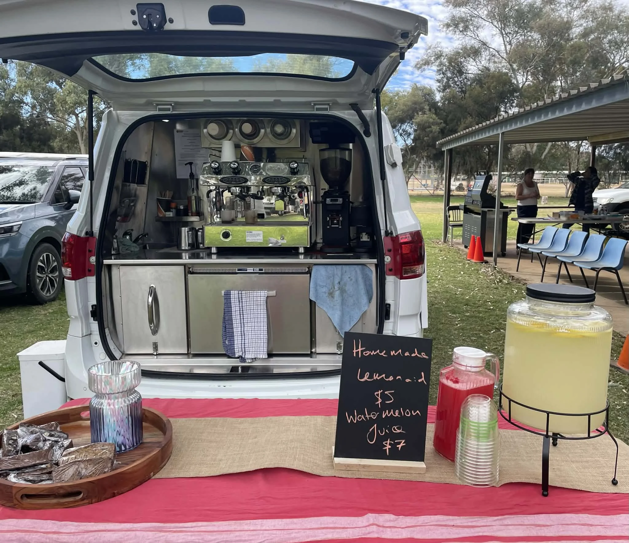 A mobile coffee stand set up at an outdoor event with a white van, serving espresso, lemonade, and watermelon juice. There is a chalkboard sign with prices, a large jar of lemonade, and a tray of baked goods on a red tablecloth.