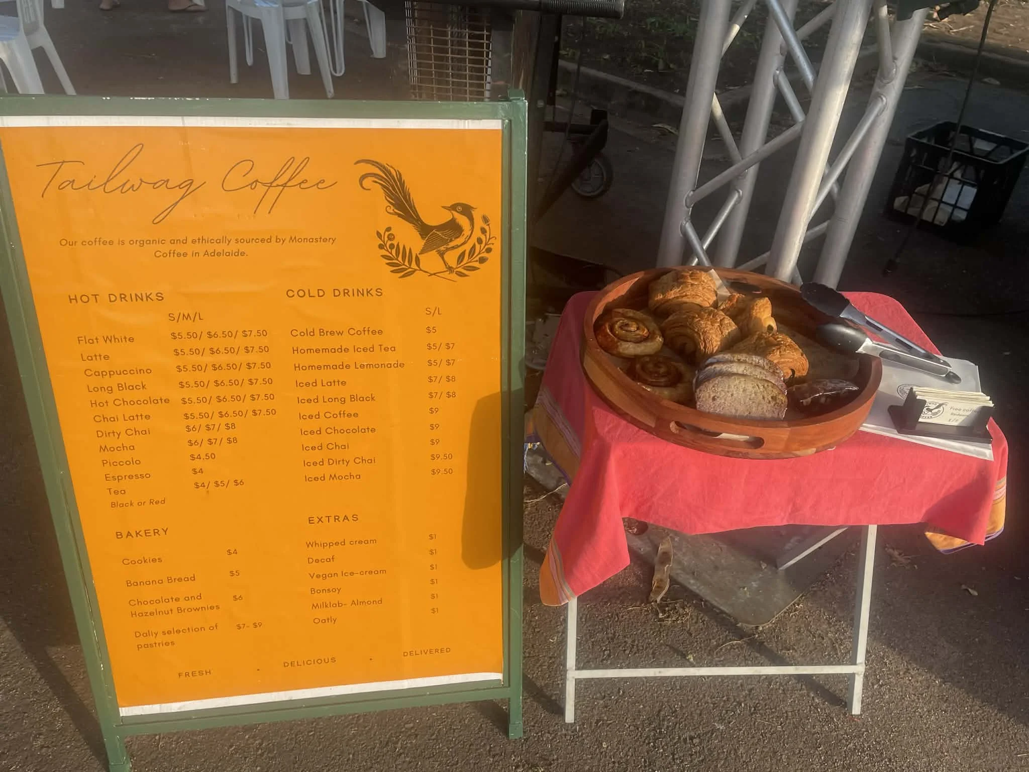 Menu board for Tailwag Coffee displaying hot and cold drinks, bakery items, extras, and prices. A table with pastry basket, tongs, and napkins next to the menu board.