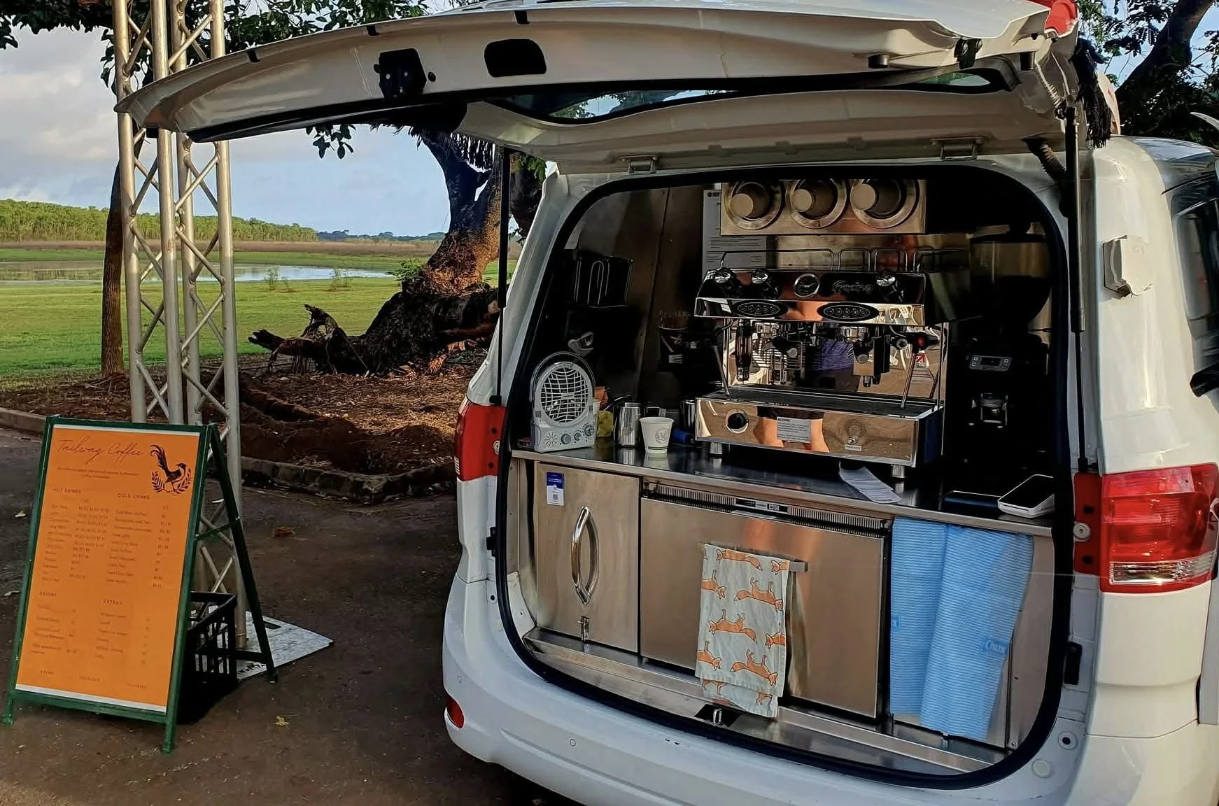 Food truck serving coffee with a menu board outside, set against a scenic outdoor background with a tree and water.