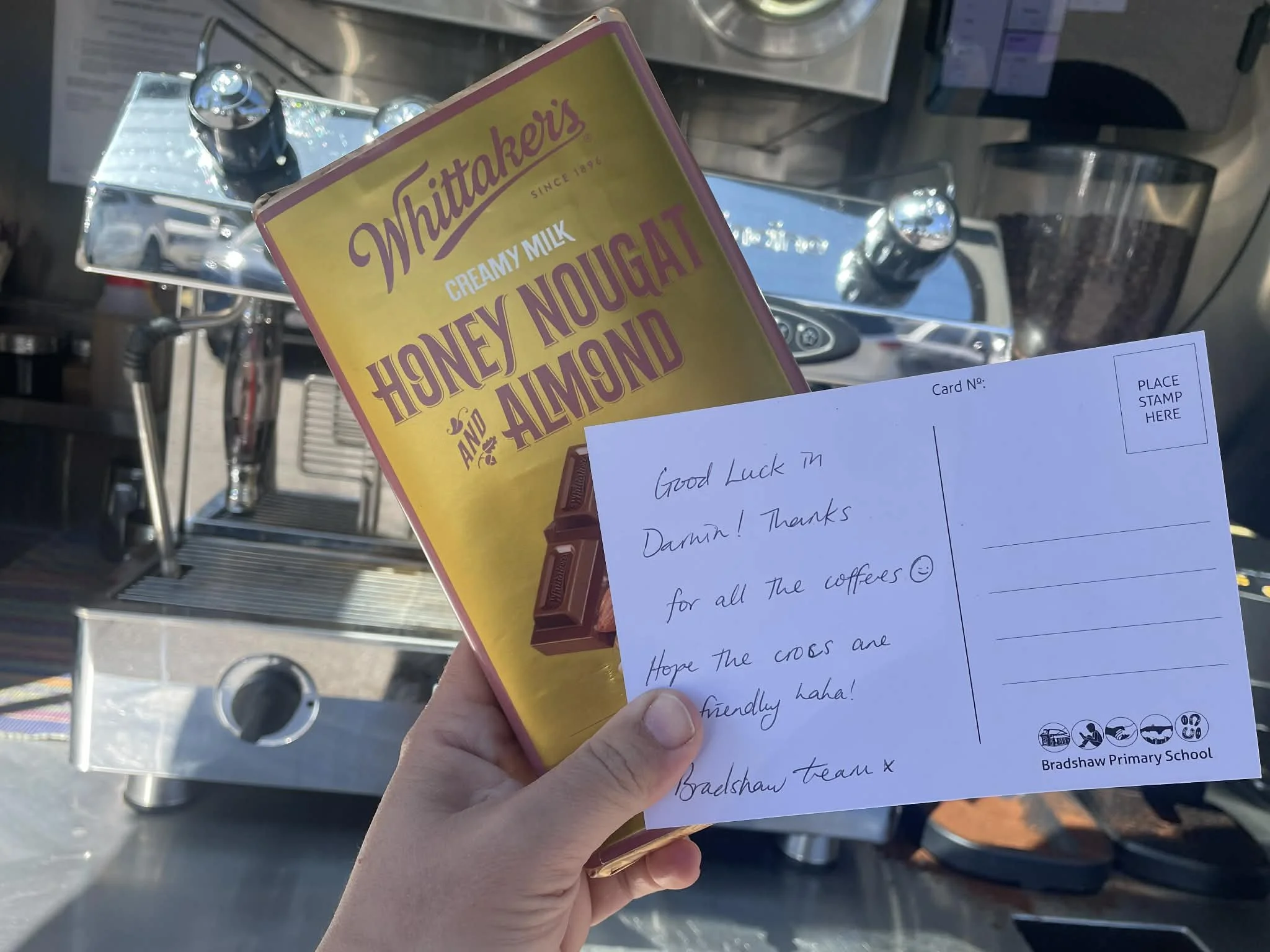 A hand holding a box of Whitaker's Honey Nougat and Almond chocolate with a handwritten note card in front of a coffee machine.