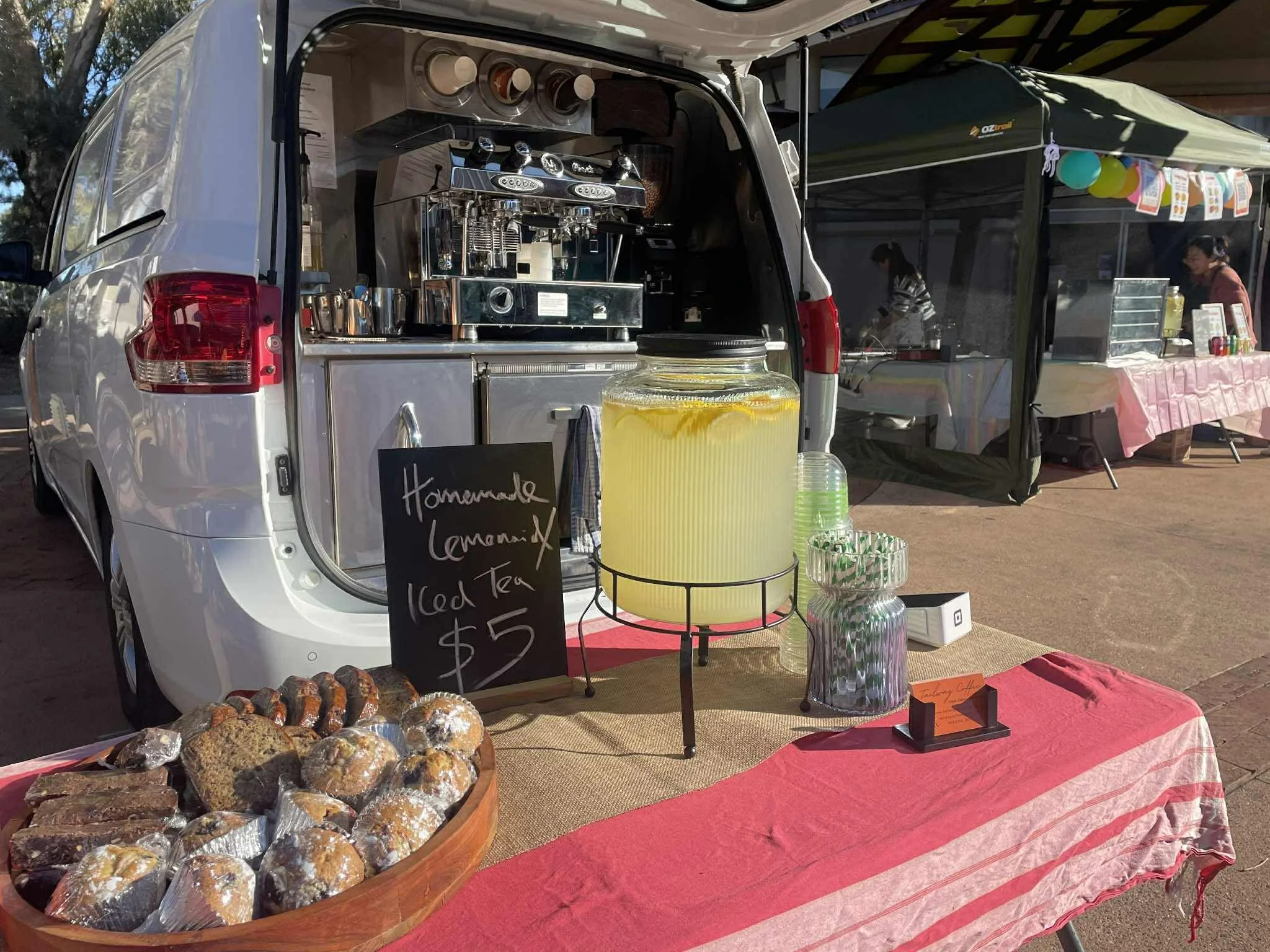 A mobile coffee and lemonade stand set up at a market. The stand features a large lemonade jar, baked goods, and a chalkboard sign advertising homemade lemonade and iced tea for $5. In the background, there are more vendor tents and people.