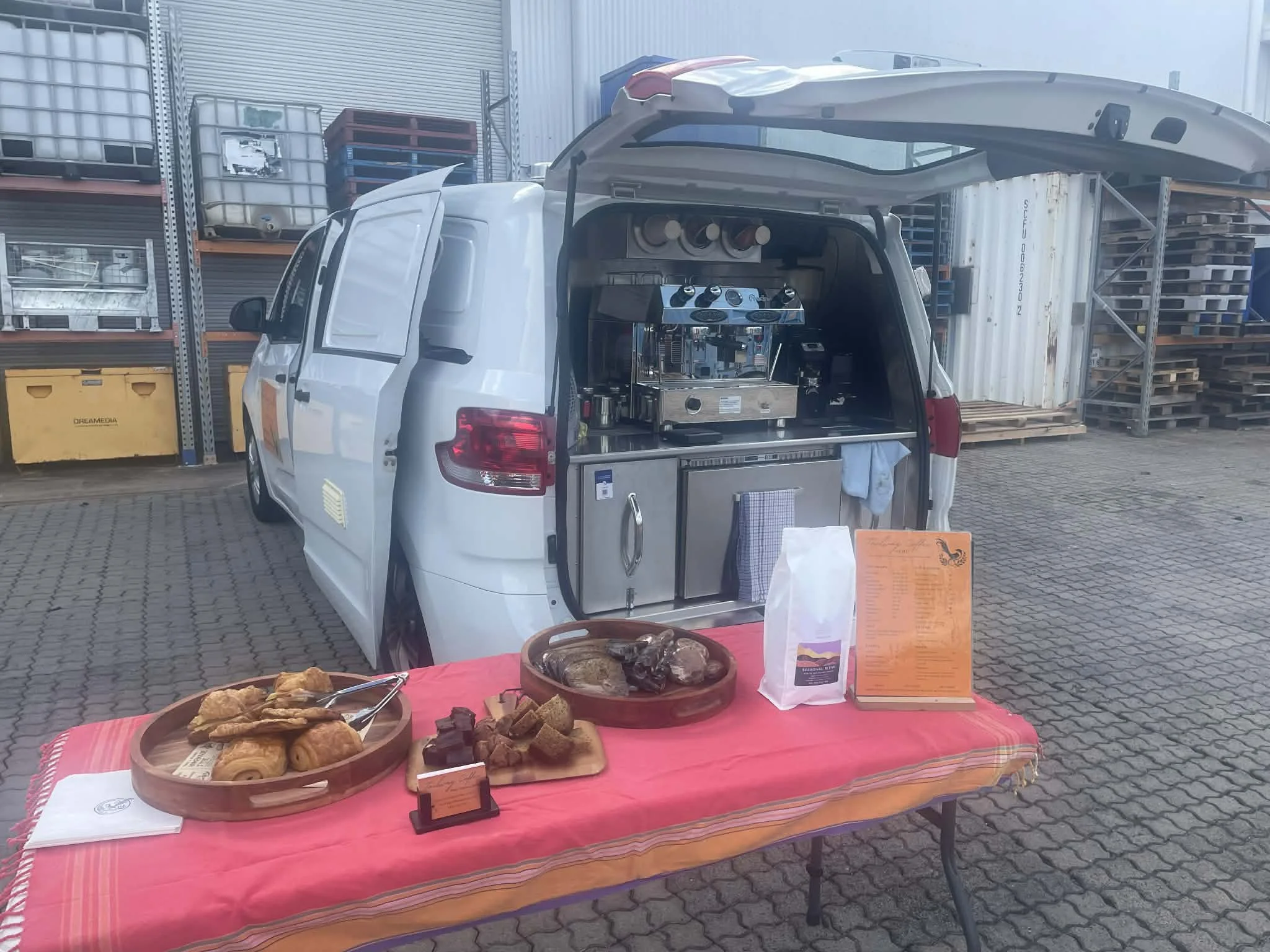 A mobile food stand set up behind a van with baked goods on a table in front. The van has its back open, revealing a coffee machine and kitchen supplies inside. The baked goods include croissants, brownies, and other pastries displayed on wooden tray