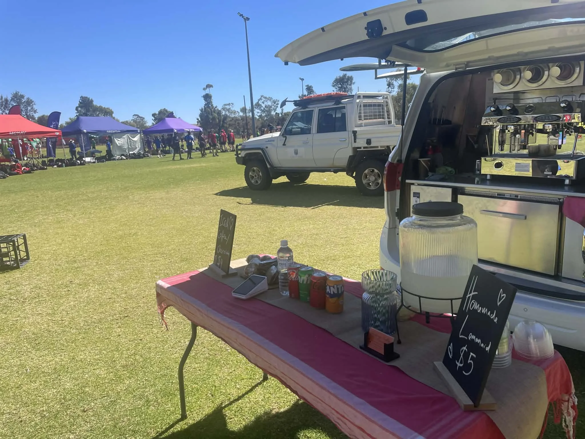 A lemonade stand setup at an outdoor event on a sunny day. The table has a pink tablecloth, bottles of drinks, a sign that reads "Homemade Lemonade $5", and a large glass jar of lemonade. Behind the table, a van with its trunk open displays coffee eq