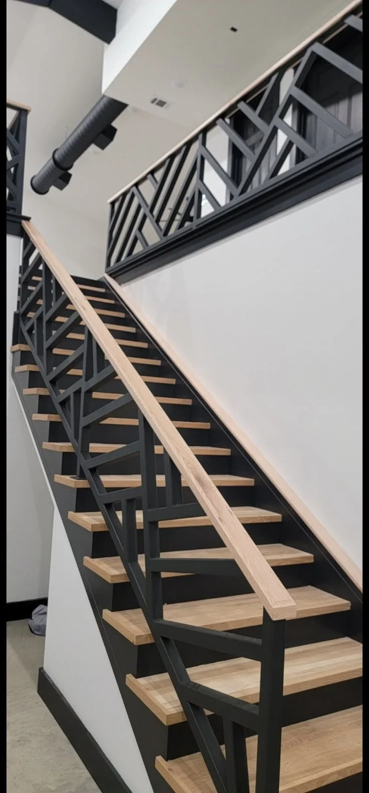 Interior view of a modern staircase with black metal framing, light wood steps, and matching handrails, in a contemporary building with white walls and black accents.