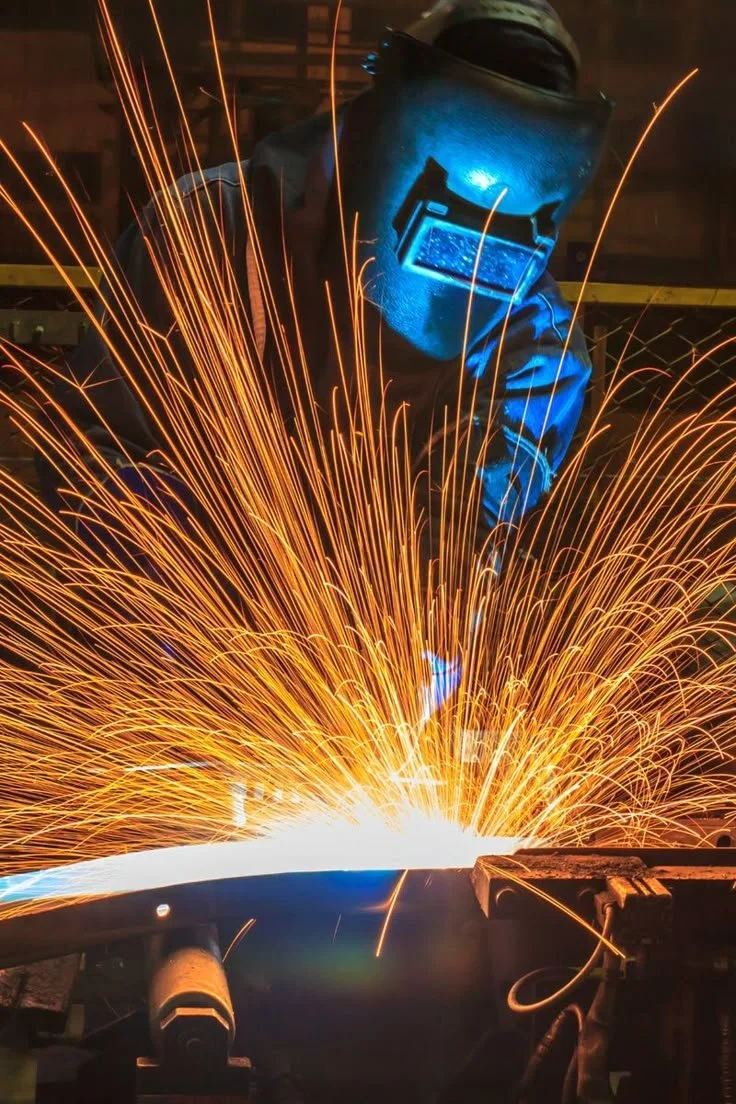 A worker wearing a welding helmet and protective gear welding metal, with bright sparks flying in all directions.