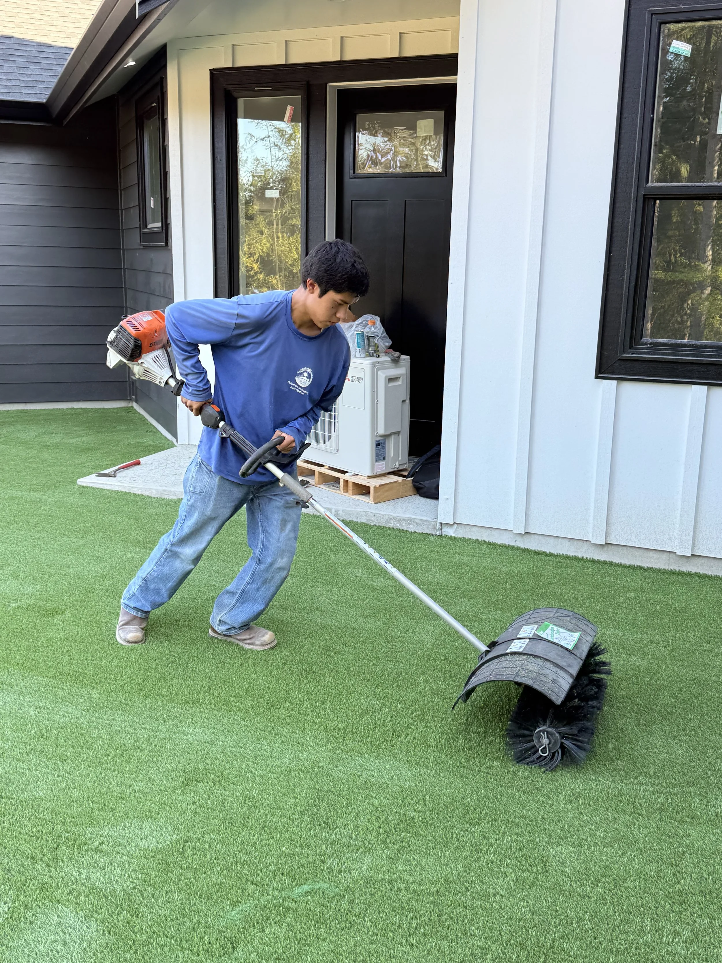 A young man is using a grass roller on a manicured lawn in front of a house with black and white siding and windows.