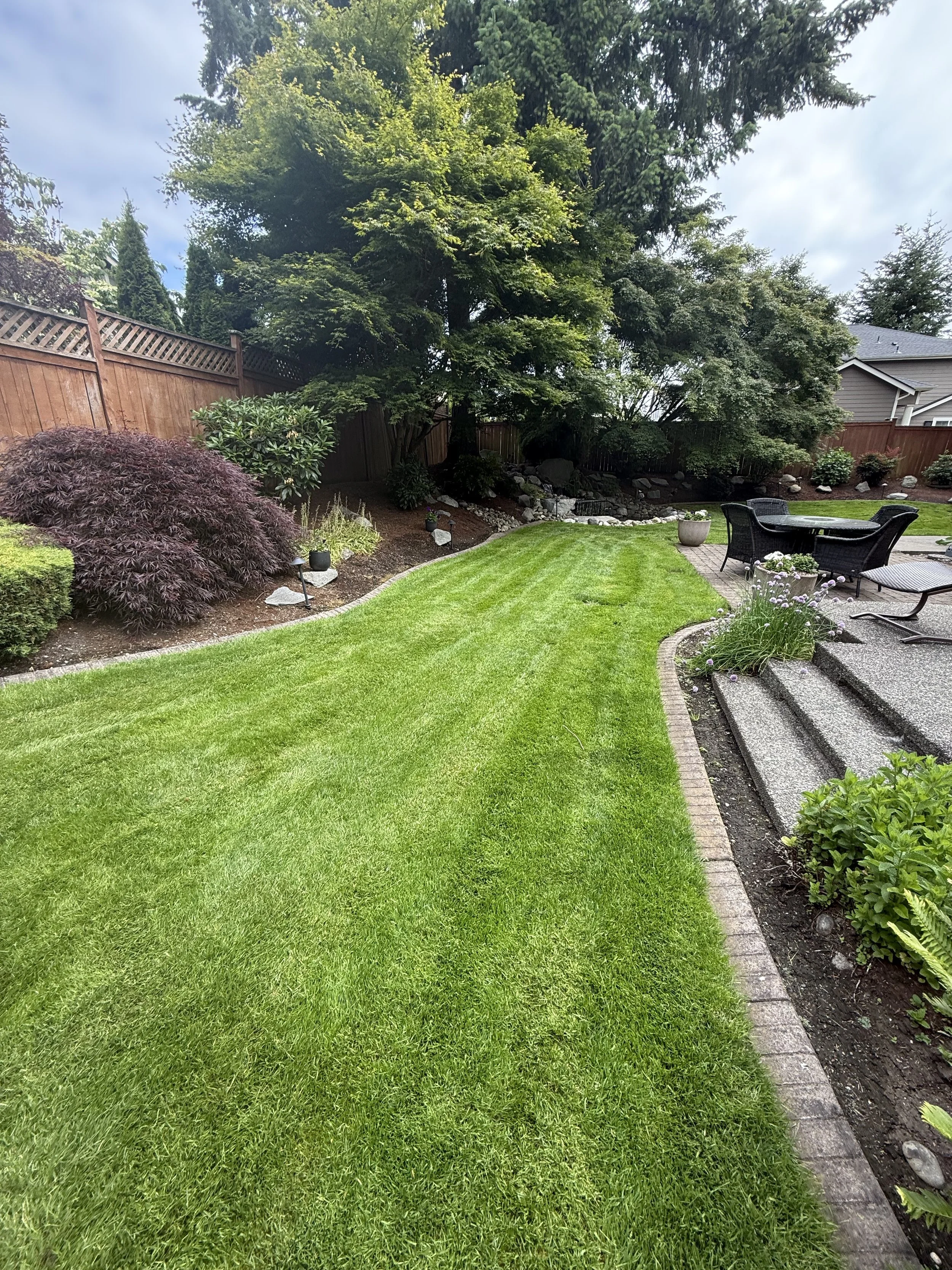 Backyard with well-manicured green lawn, garden beds with shrubs and small trees, a patio with outdoor furniture, and a wooden fence under a partly cloudy sky.