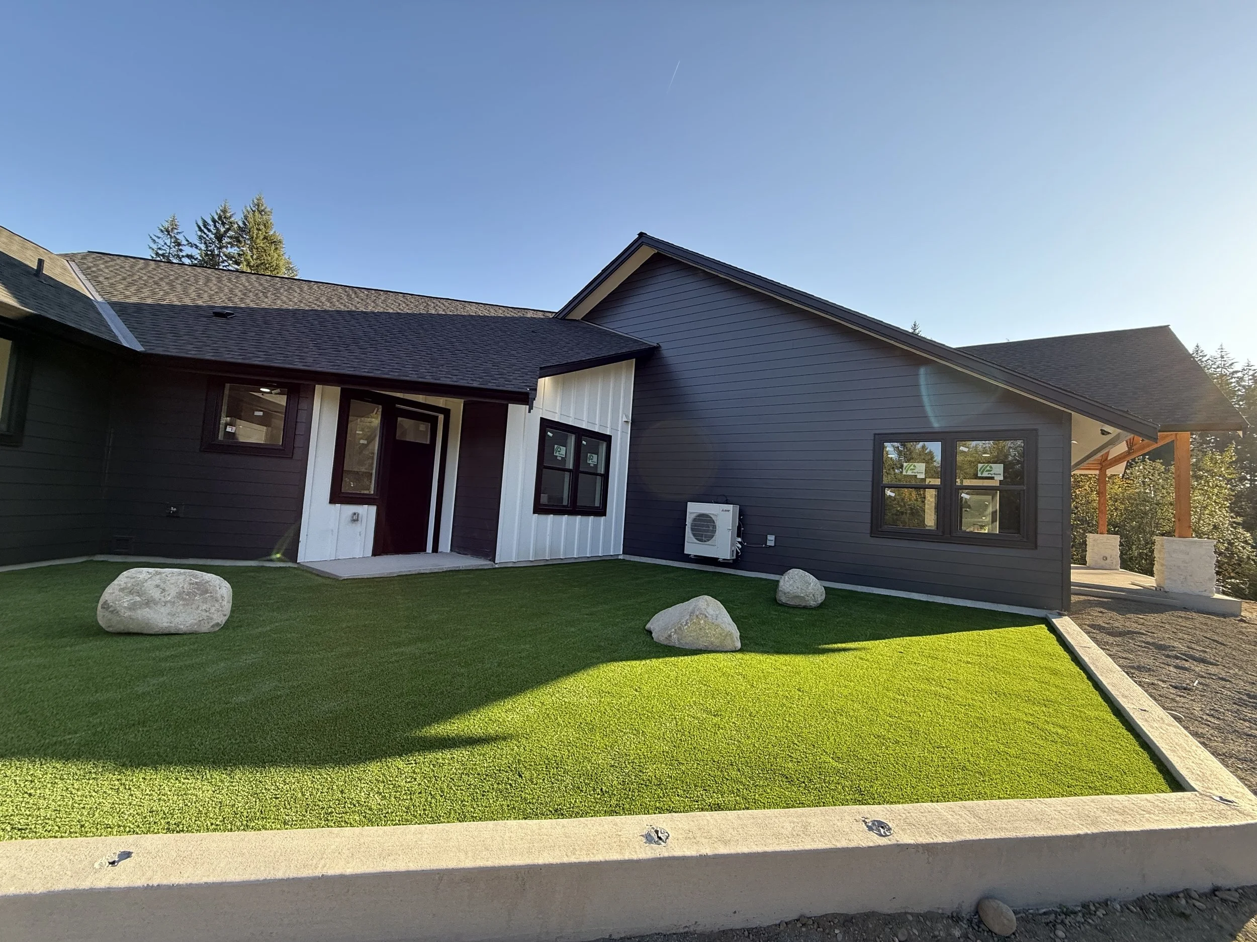 New modern house with dark gray and white exterior, front yard with artificial grass and large rocks, clear blue sky.