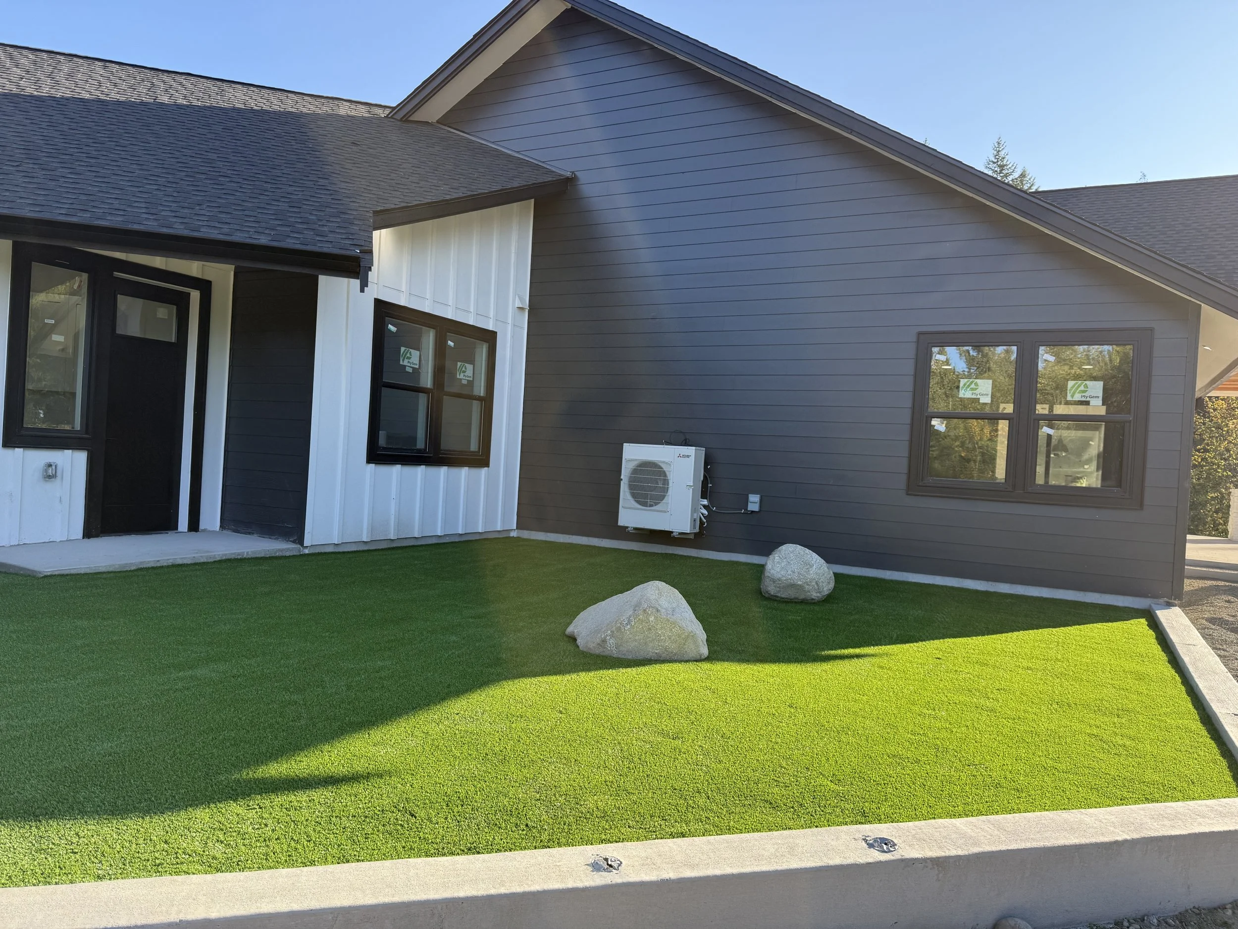 A modern house with dark gray and white siding, a manicured green lawn, and an outdoor AC unit mounted on the wall. There are three large rocks on the lawn and a concrete pathway near the entrance.