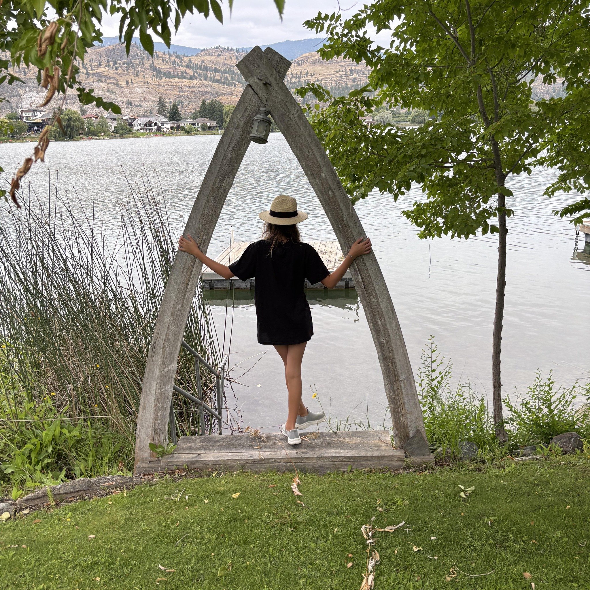 A woman wearing a black dress, white sneakers, and a beige sunhat stands on a wooden platform at the edge of a lake, holding onto a wooden archway, looking out at the water and distant hills.