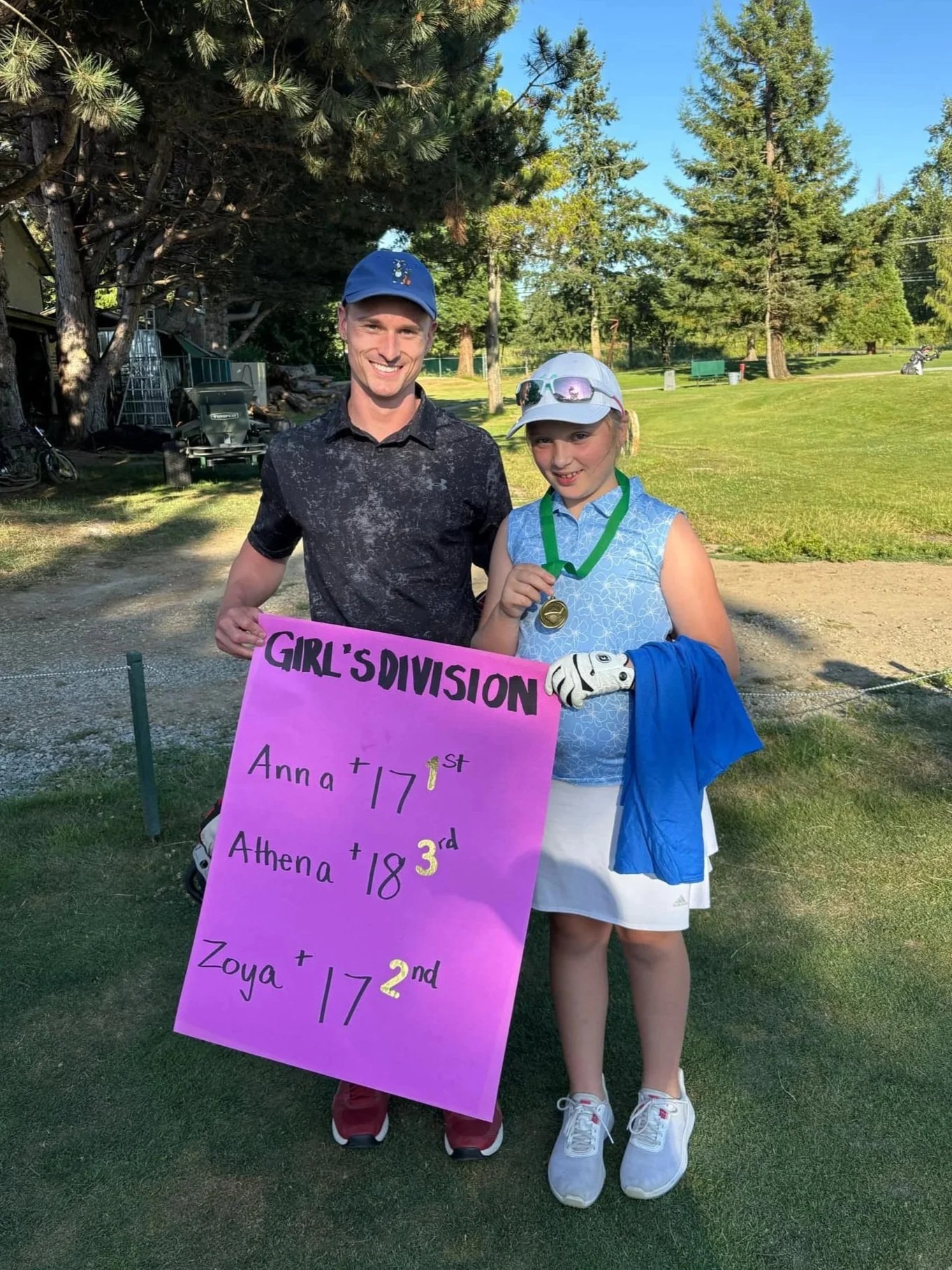 A young girl and a man are standing outdoors on a grassy area with tall trees in the background. The girl is wearing a white cap, sunglasses, a light blue sleeveless top, white skirt, and white shoes, holding a gold medal. She has a blue jacket draped over her arm. The man is wearing a cap, a black patterned polo shirt, and red pants. They are smiling, and the girl is holding a purple sign with black and yellow writing that lists names and rankings for a girls' golf division, indicating the girl as second-place winner. The setting suggests a golf course or park during daytime.