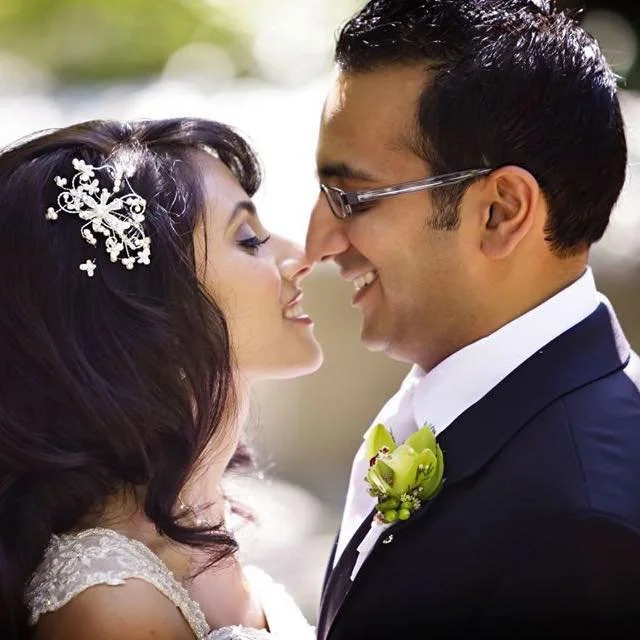 A bride and groom are facing each other close, smiling with their noses almost touching outdoors. The bride has dark hair with a decorative white hairpiece, and the groom is wearing glasses and a dark suit with a white shirt and a boutonniere.