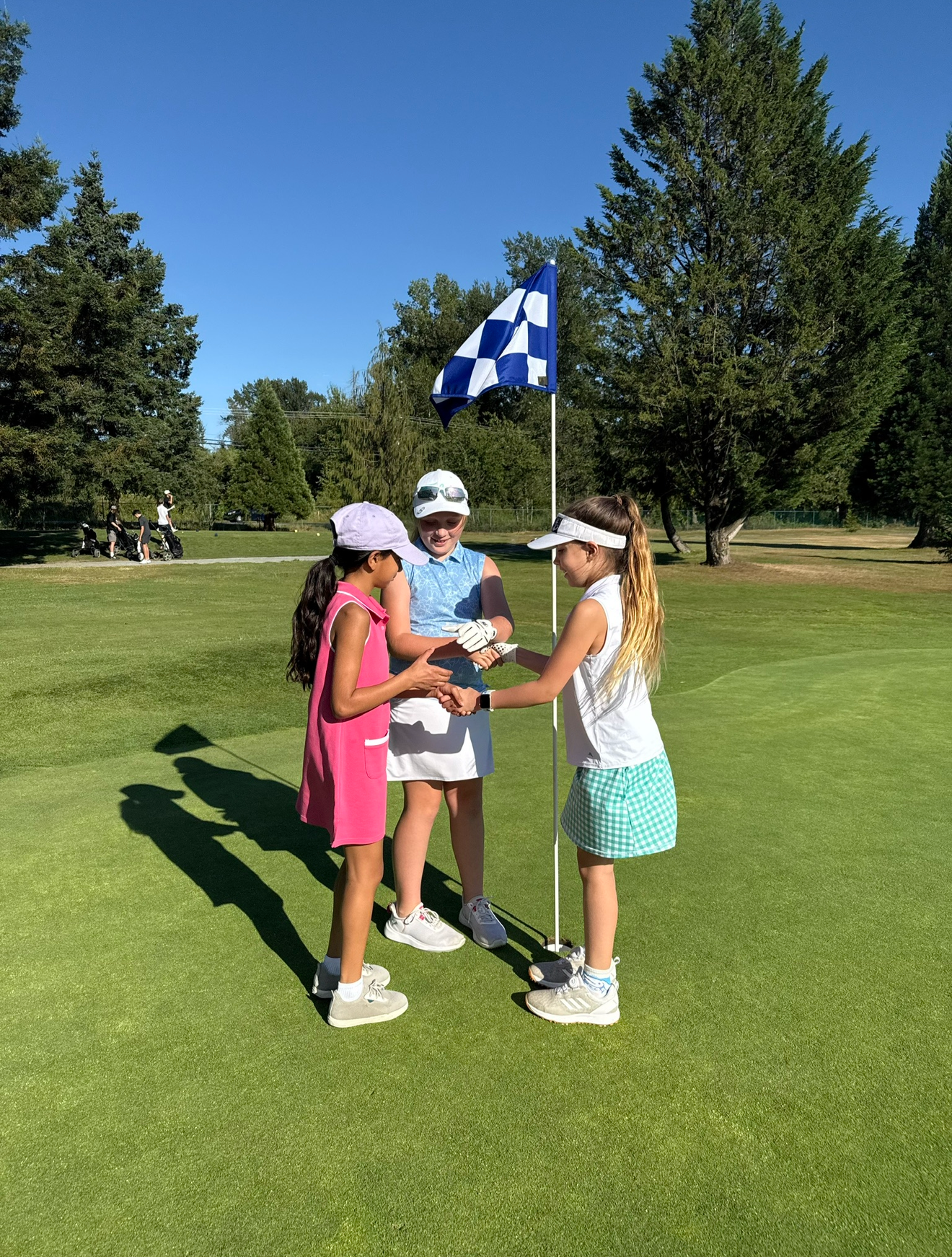 Three young girls on a golf course participate in a golf award ceremony, with two of them shaking hands while holding a plaque, under a blue and white checkered flag on a sunny day.