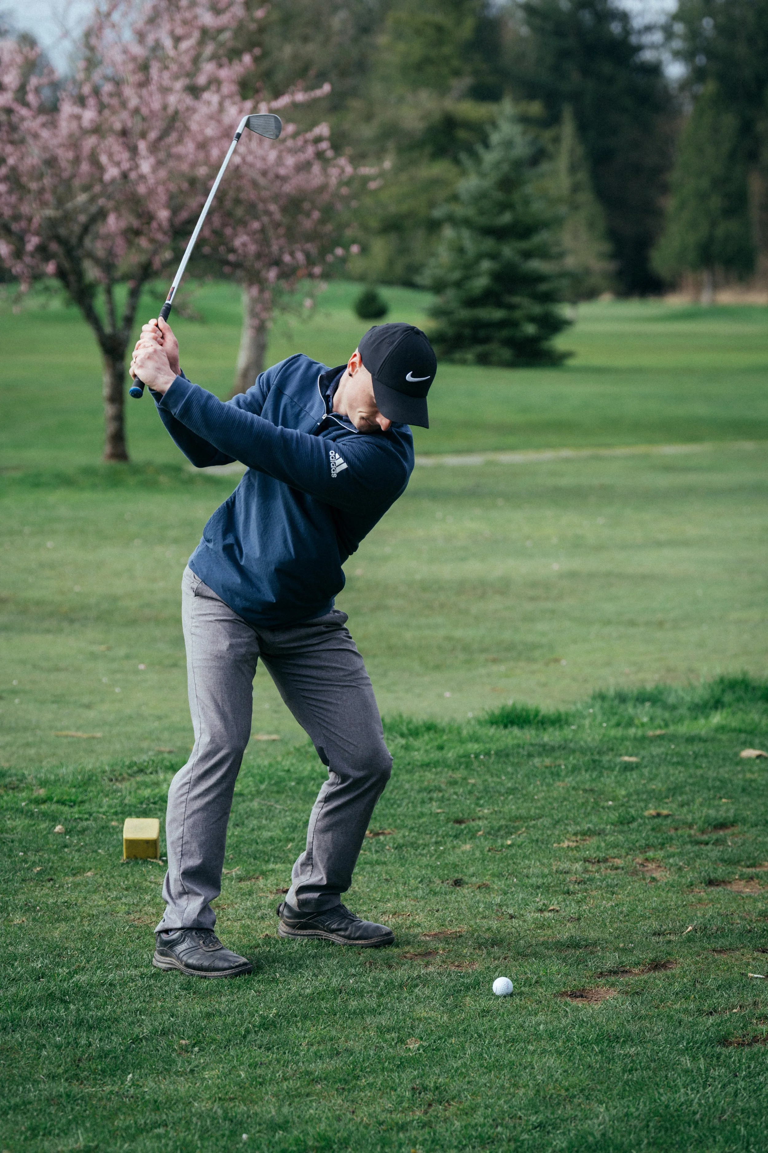 A man in a navy jacket and black cap is swinging a golf club on a golf course with green grass and trees in the background.
