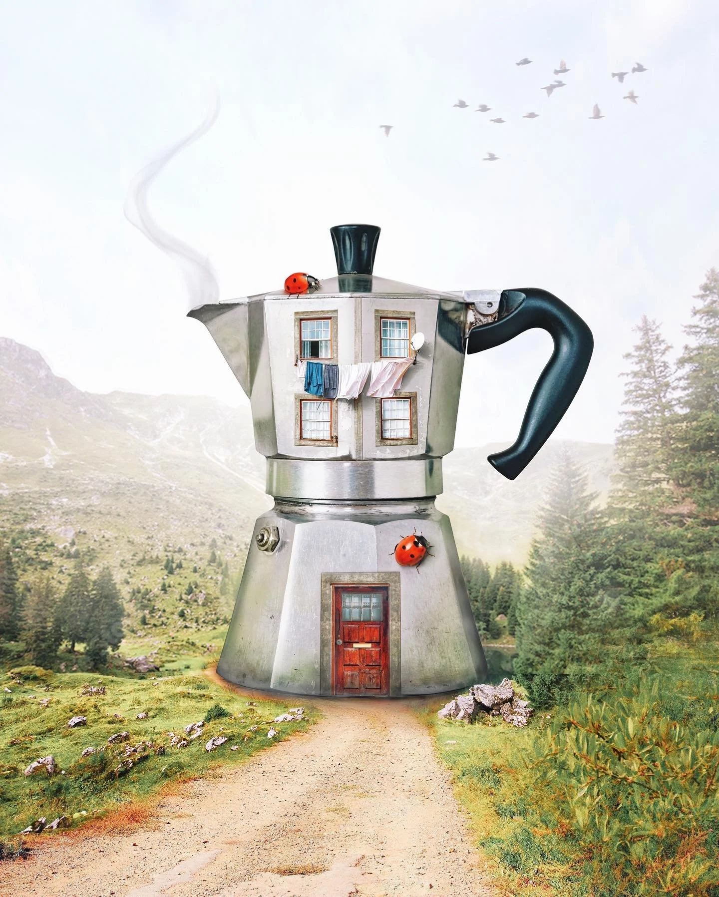 A large coffee maker with a house design on its upper part, situated on a dirt path in a scenic outdoor setting with mountains and trees. The house features windows with clothes hanging outside, and the coffee maker has a black handle, red ladybug decorations, and a chimney emitting smoke.
