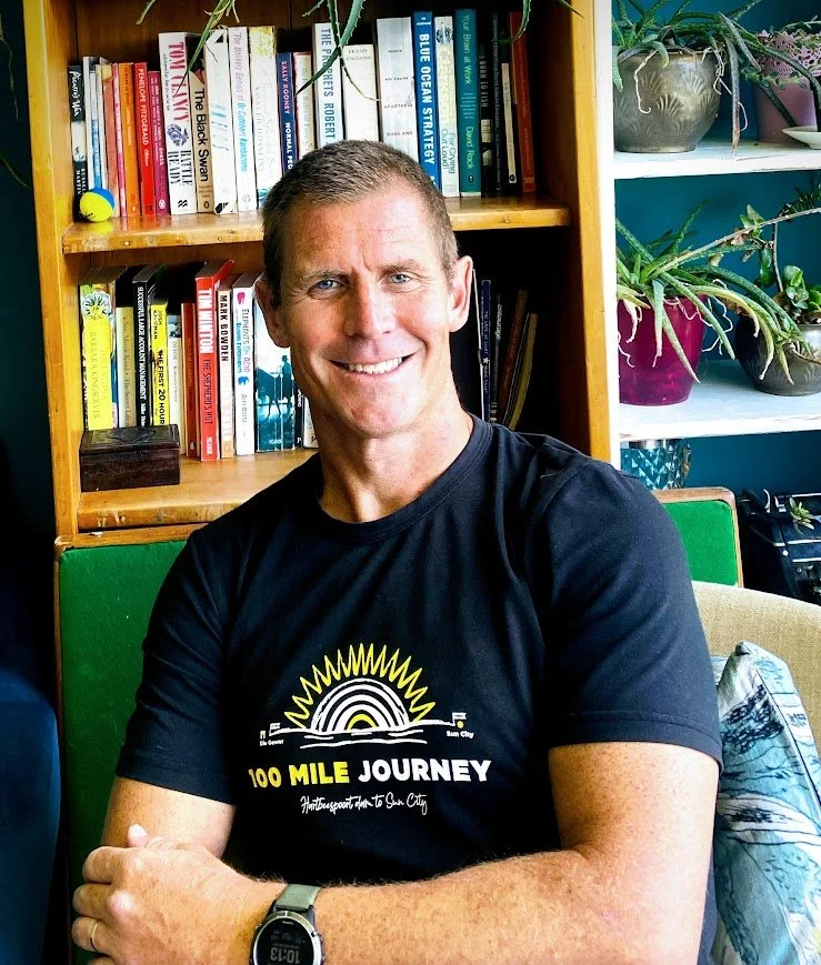 Smiling man in black T-shirt sitting in front of bookshelf with books and plants.