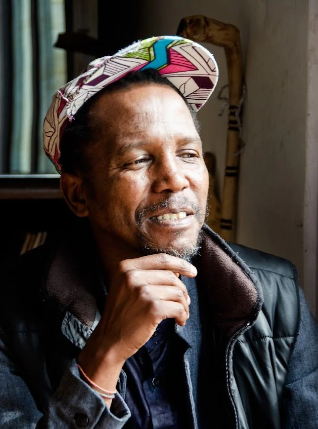 A man with a beard and mustache wearing a colorful patterned cap, a dark jacket, and a gray shirt, smiling thoughtfully while resting his chin on his hand indoors.