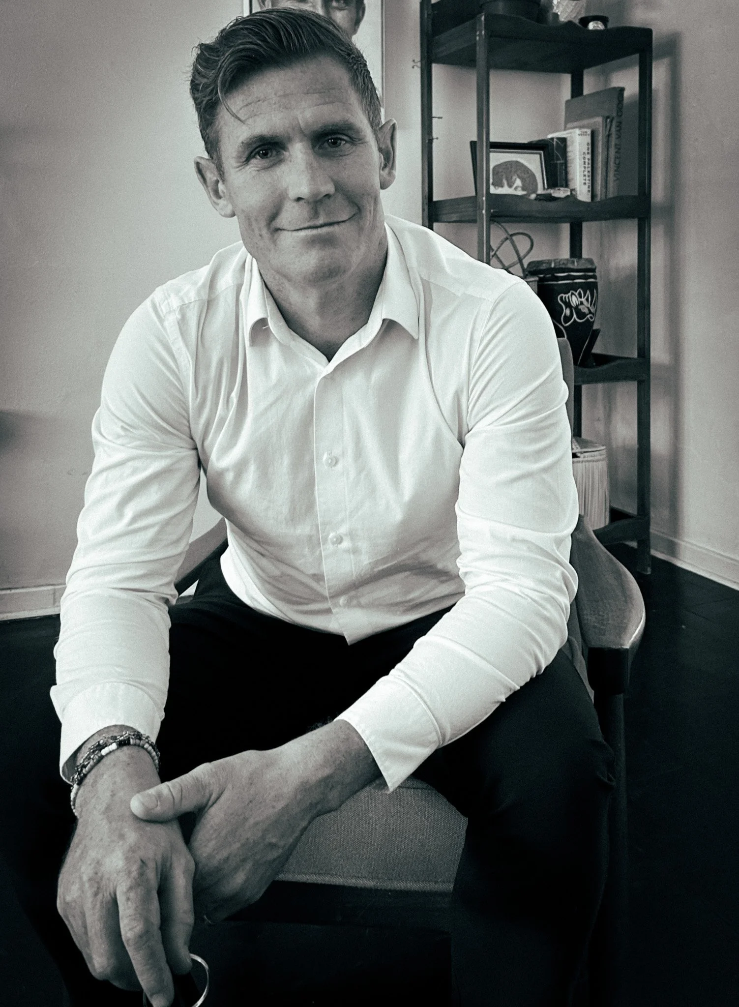 Smiling man in black T-shirt sitting in front of bookshelf with books and plants.