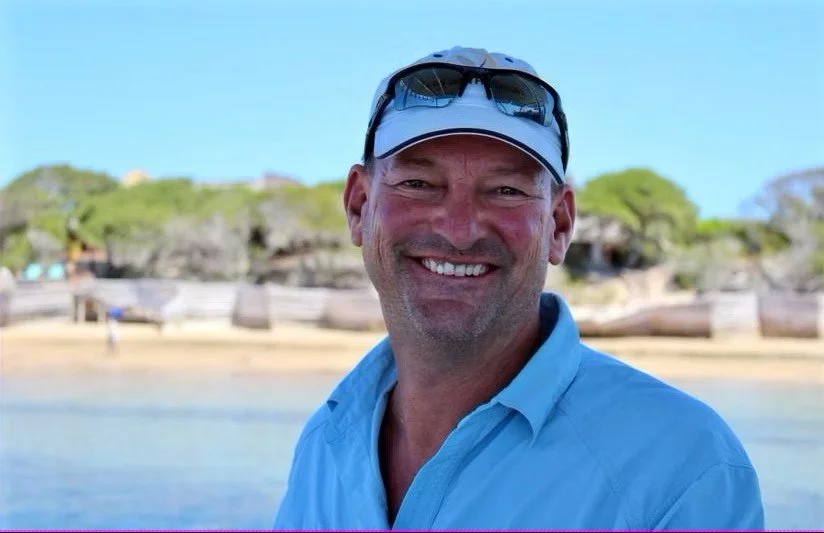 Smiling man in a blue shirt wearing sunglasses and a cap, standing outdoors near water with trees and a sandy area in the background.