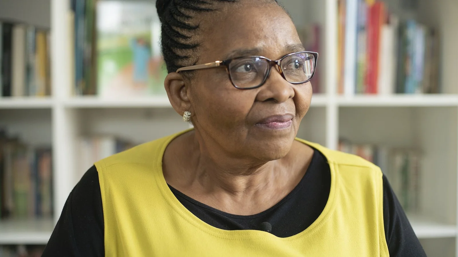 An older Black woman with glasses and earrings wearing a yellow vest sitting in front of a bookshelf.