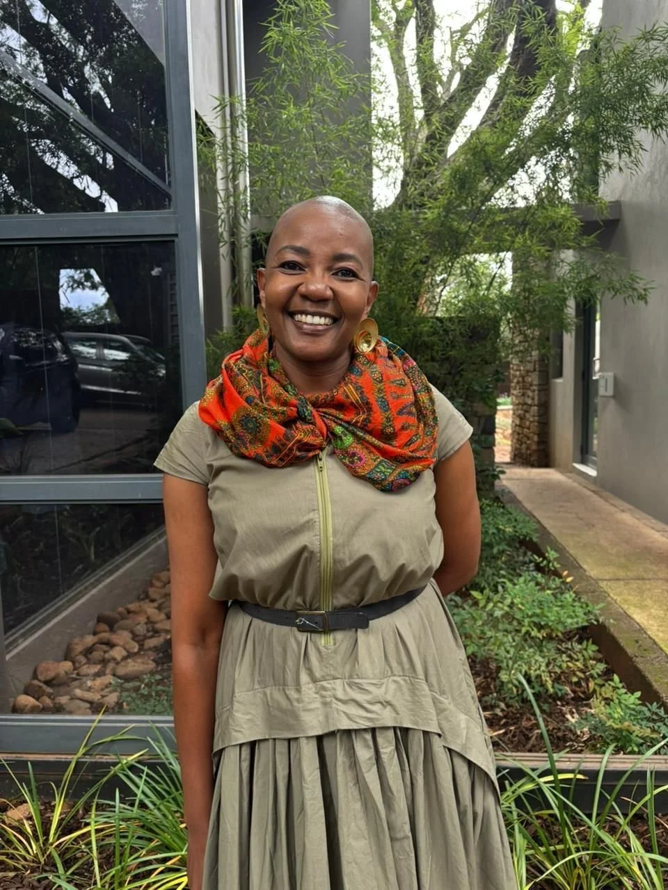 Smiling woman with a shaved head wearing a beige dress with a colorful scarf around her neck, standing outdoors in front of a garden with trees and plants.