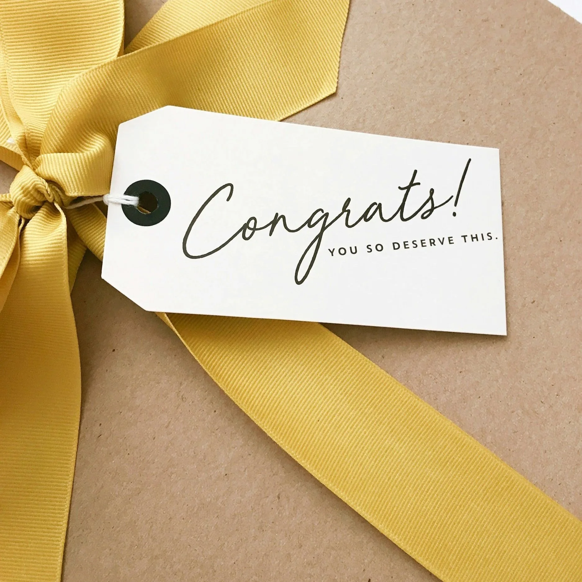 Close-up of a yellow ribbon tied in a bow alongside a gift tag that says 'Congratulations! You so deserve this.' on brown paper.