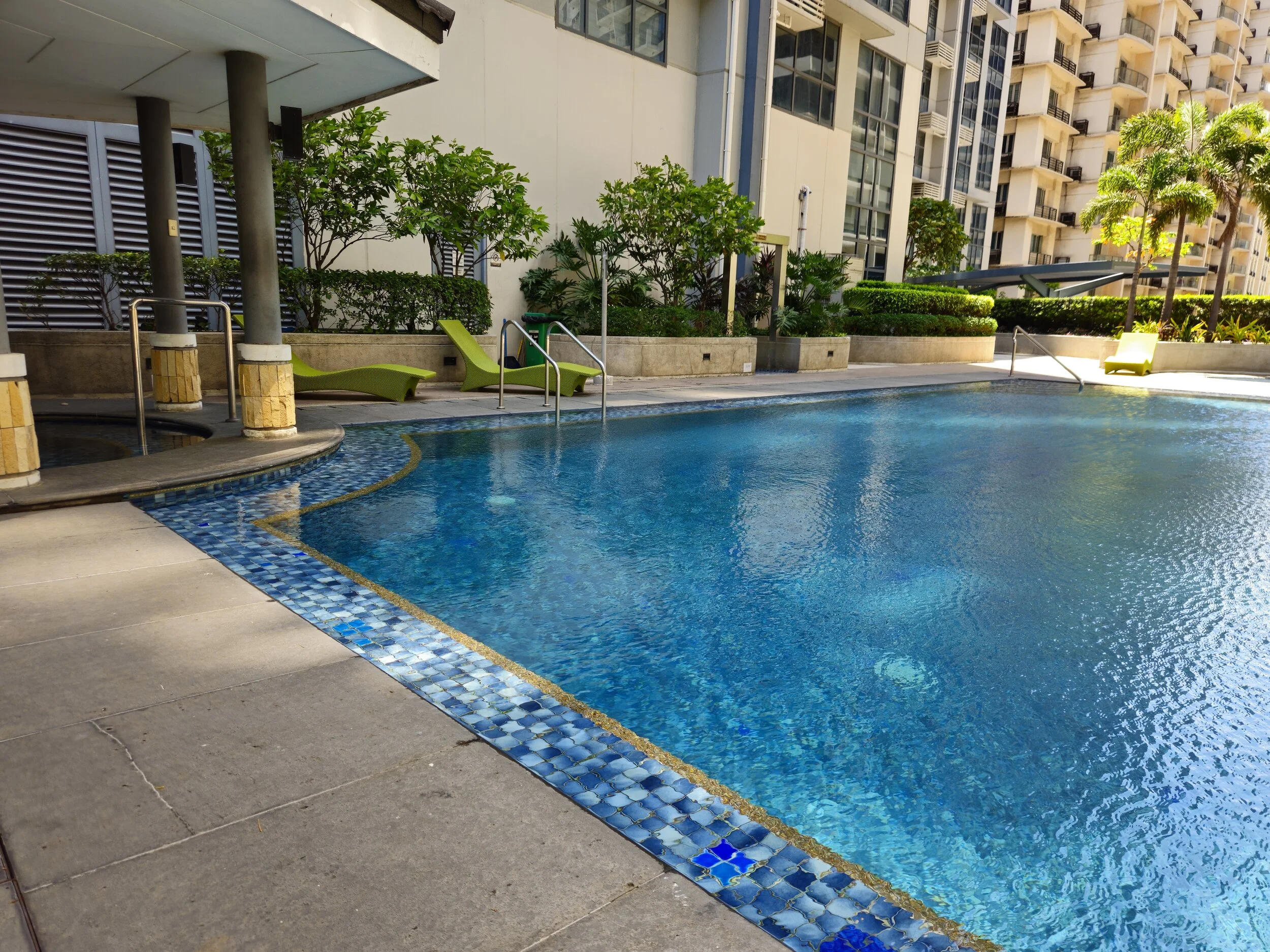 Outdoor swimming pool with blue water, surrounded by lounge chairs and greenery, in a modern residential building complex.