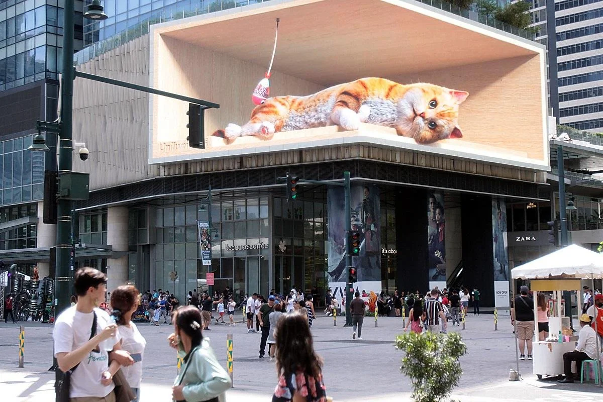A massive 3D billboard of a playful, orange tabby cat in a wooden box captivates pedestrians on a busy city street. The mood is lively and curious.