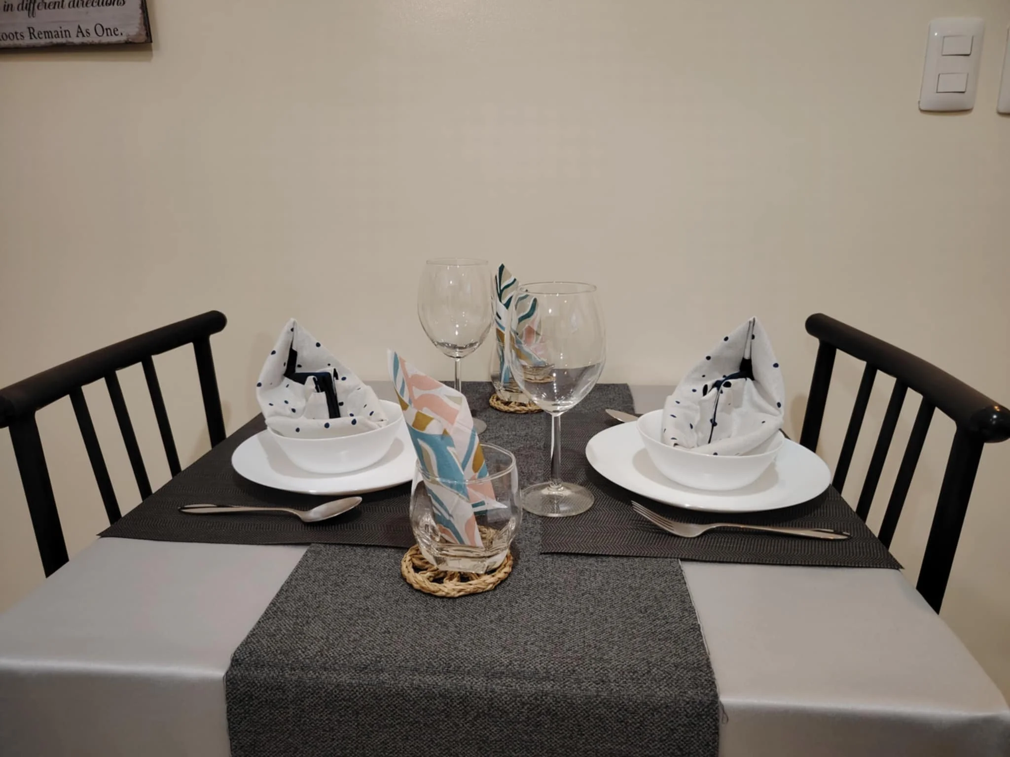 A cozy dining table set for two with white plates, folded napkins, wine glasses, and cutlery on a gray tablecloth. The setting is elegant and inviting.