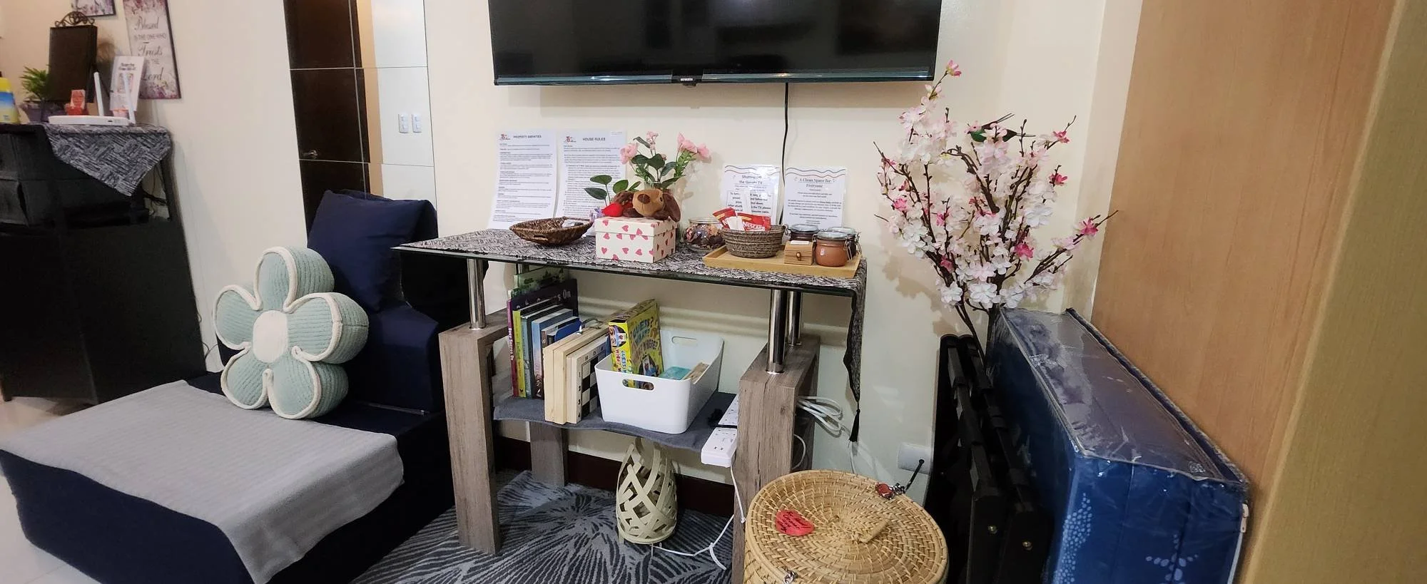 A cozy living room features a TV above a decorated console table with pink flowers and books. A blue chair with a floral pillow adds comfort.