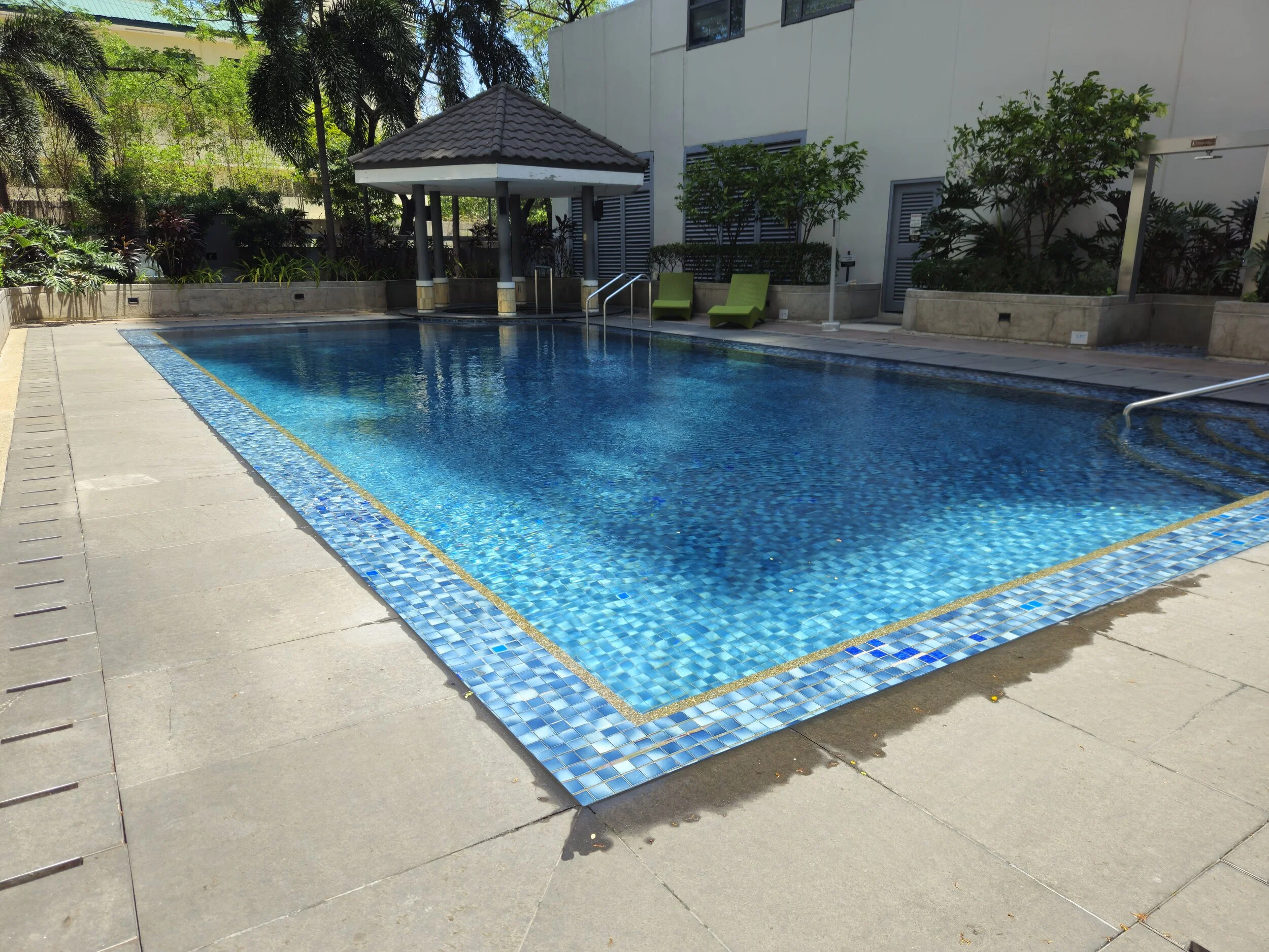 Outdoor swimming pool with blue tiles, surrounded by a concrete deck, with two green lounge chairs and a gazebo in the background.