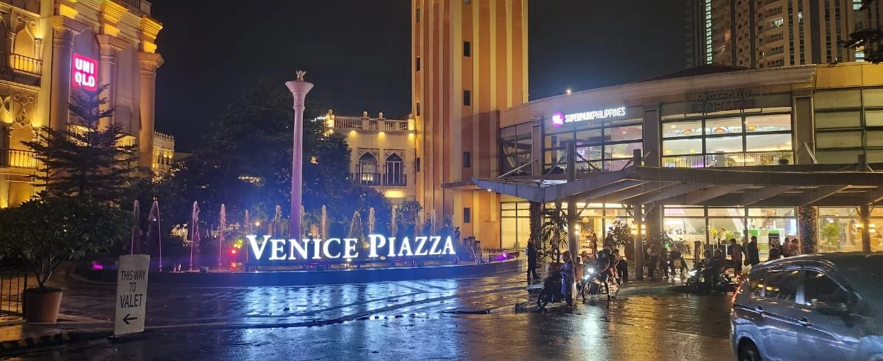 Night time view of Venice Piazza, illuminated with glowing fountains and surrounding buildings. A bustling scene with people gathered, conveying a lively ambiance.