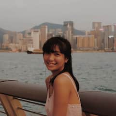 A young girl with black hair smiling at the camera, standing near water with a city skyline backdrop.