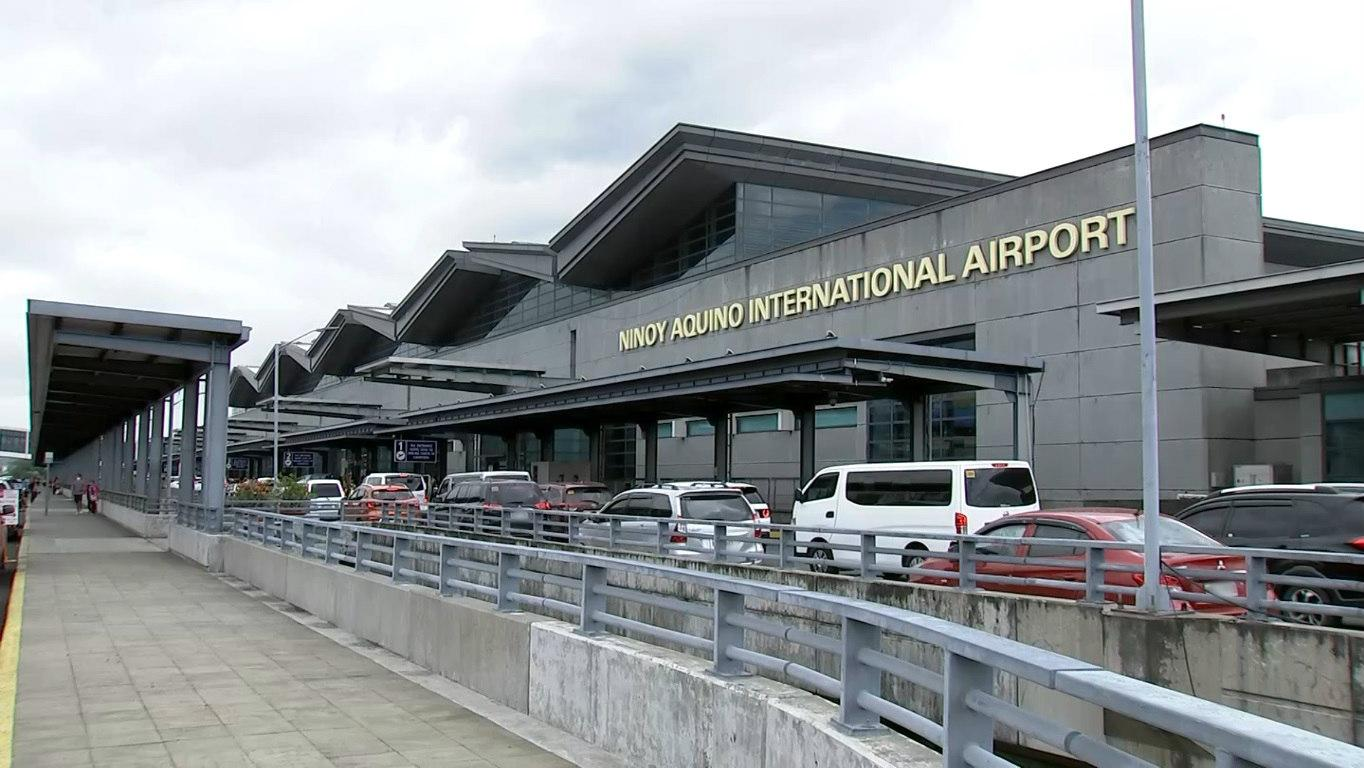 Exterior view of an airport terminal with large, modern architecture. Several cars are parked in front, and cloudy skies loom above, creating a busy yet calm atmosphere.