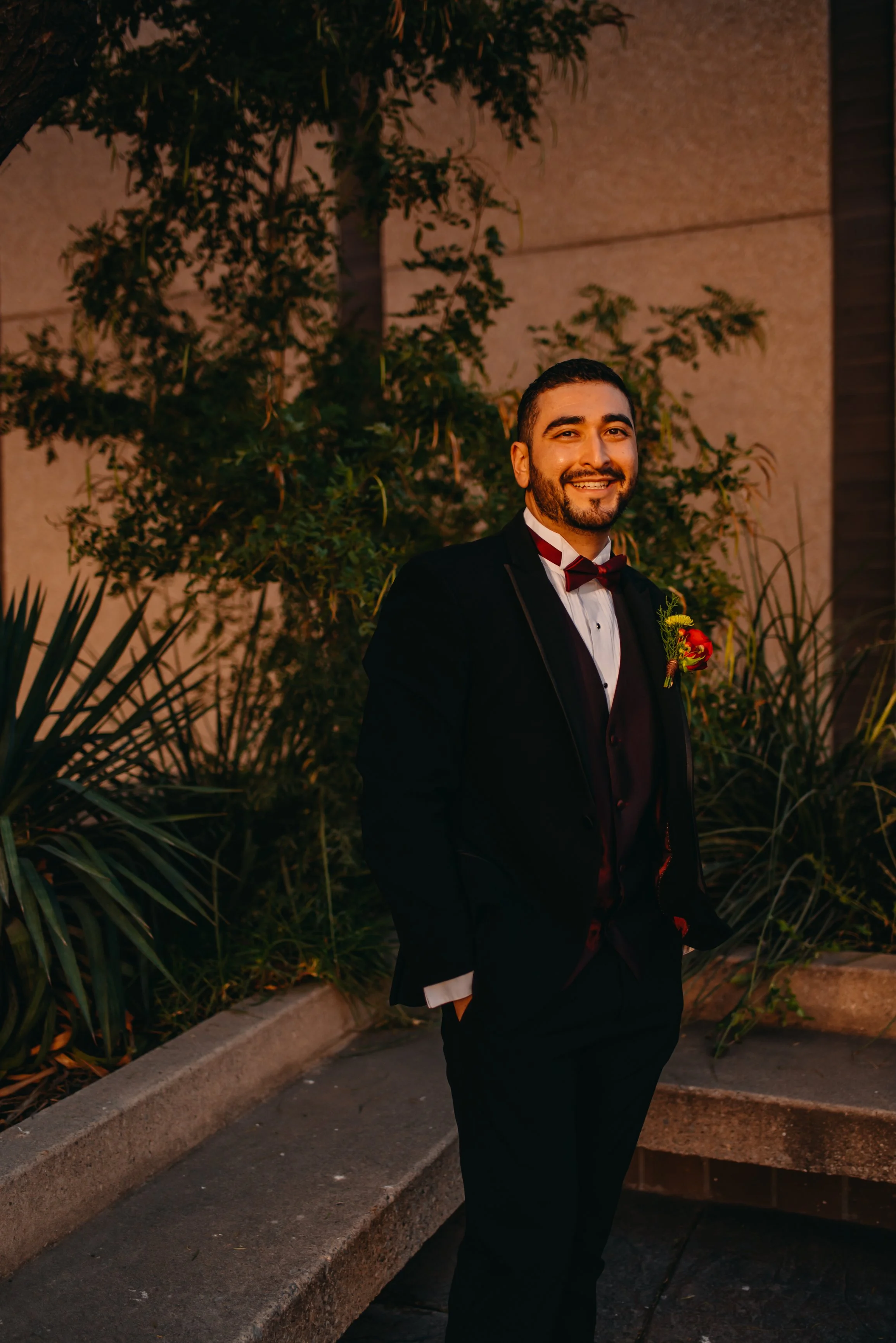 A man in a black tuxedo with a maroon bow tie and boutonnière stands outdoors at night in front of greenery and a beige stone wall, smiling at the camera.