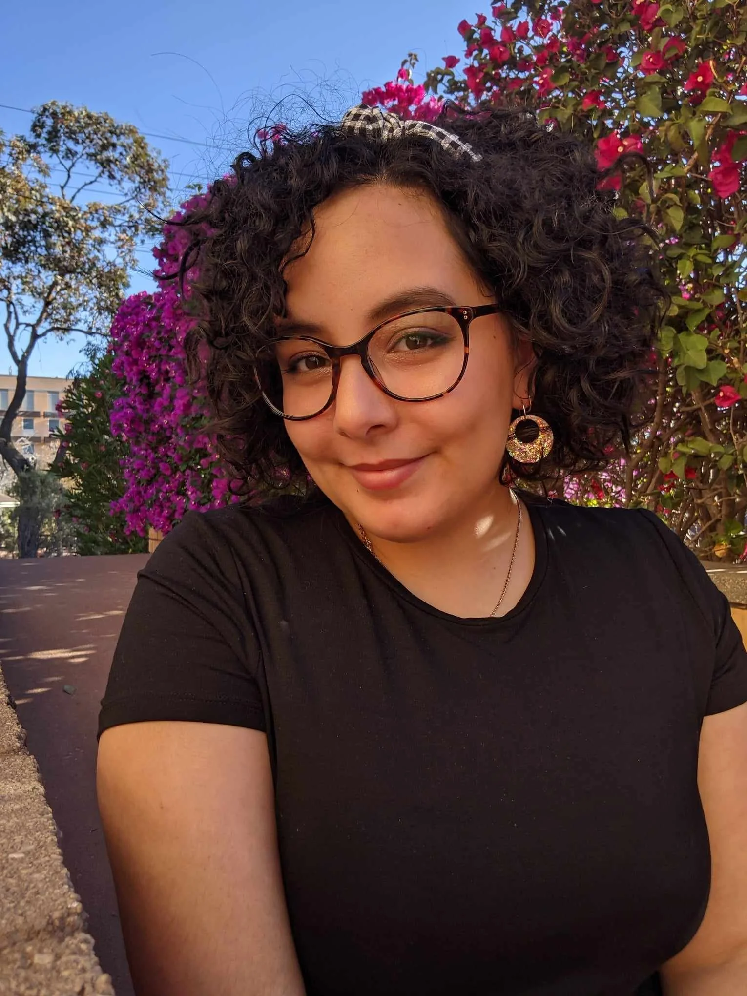 A young woman with curly dark hair and glasses, smiling outdoors with pink bougainvillea flowers and blue sky in the background.