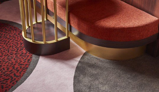 Close-up of a modern seating area with a rounded red bench. The design includes a gold railing and circular patterns on a multicolored carpet featuring red, gray, and black hues.