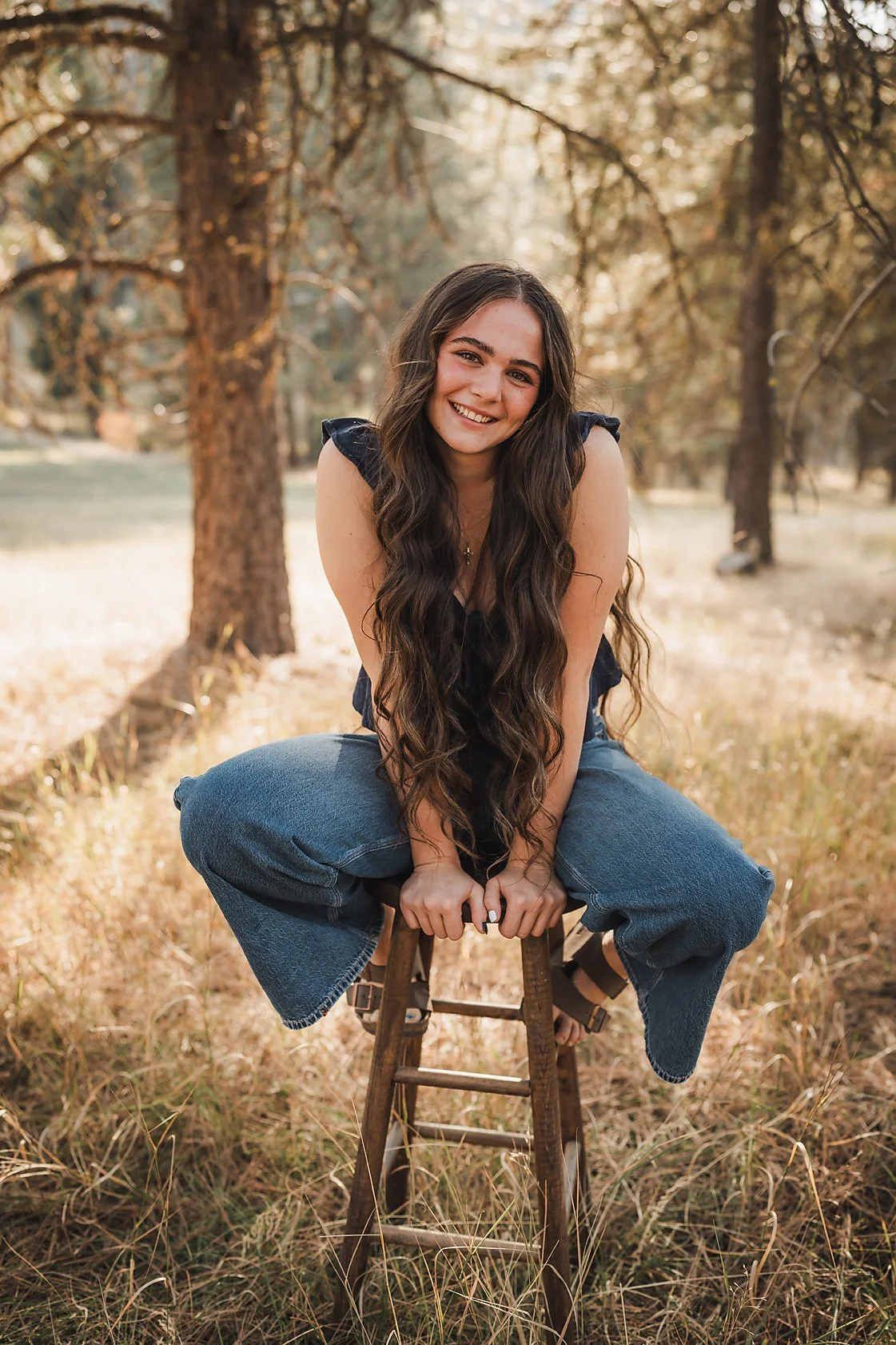 Eagle High School senior girl smiling on a wooden stool in a sunlit Garden Valley Idaho pine forest during her senior session