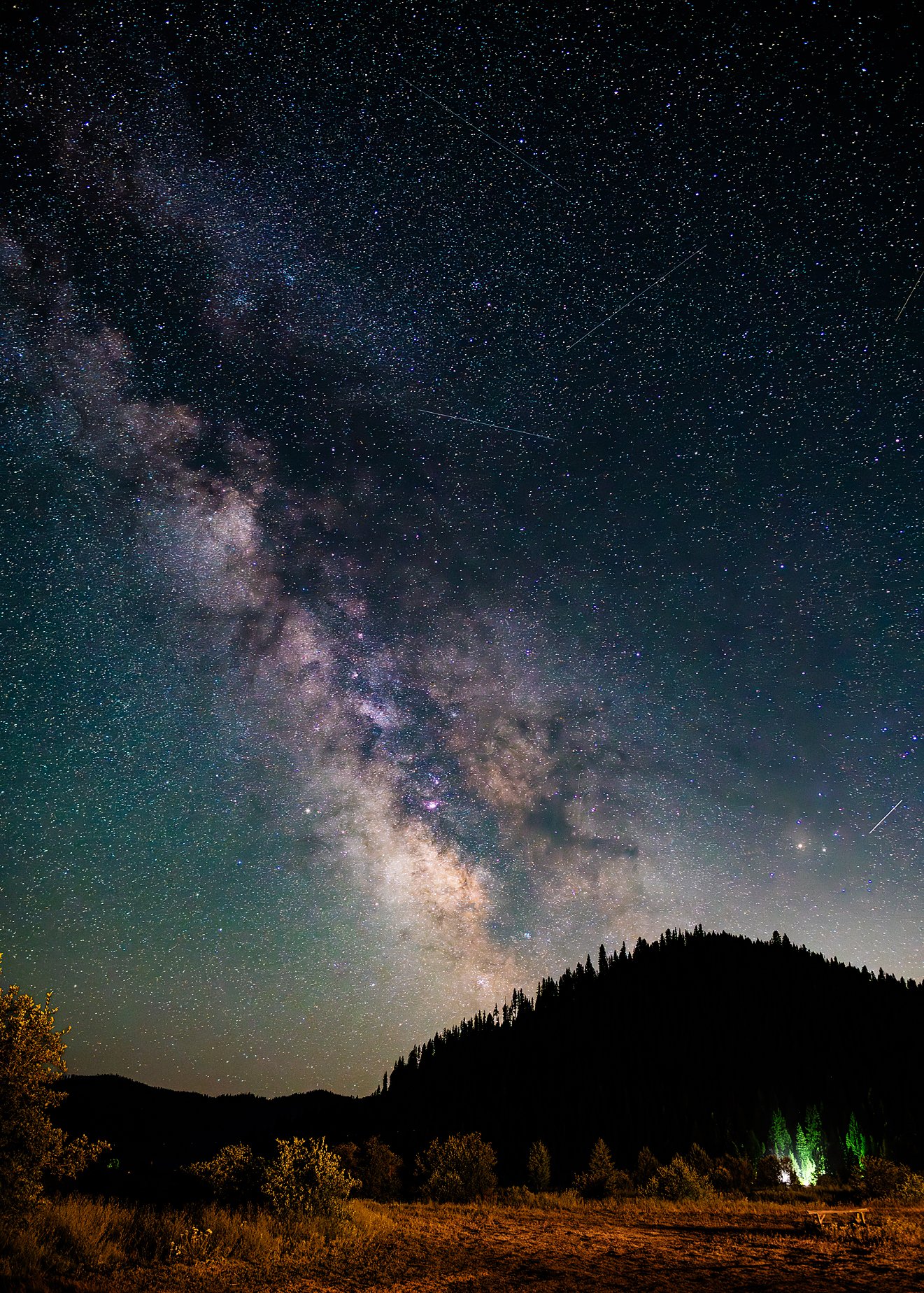 Milky Way and meteors over forested Idaho mountain with distant headlights through trees — astrophotography by Kim Starkey