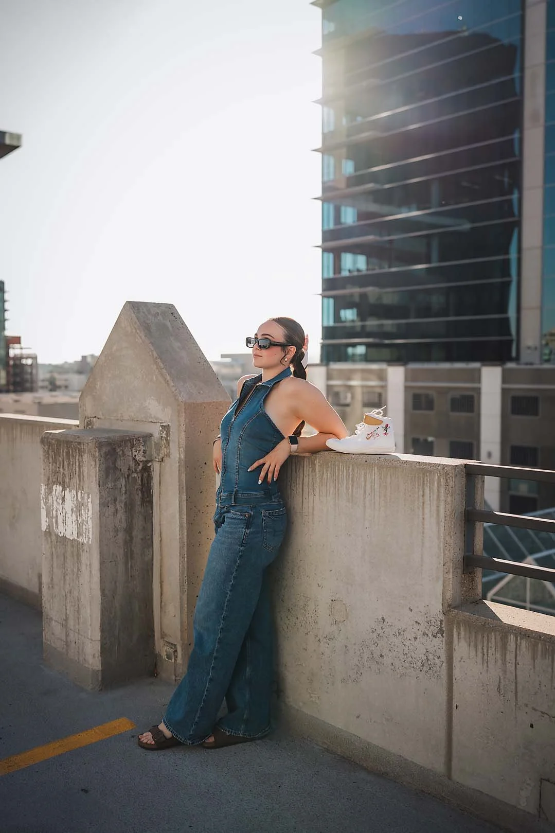 Eagle High School senior wrestler in denim and sunglasses leaning on a rooftop ledge next to her wrestling shoes with downtown Boise Idaho buildings behind her