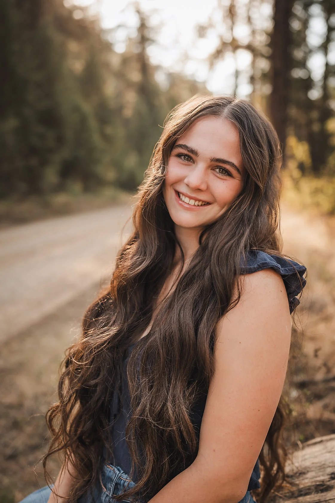 High school senior girl with long wavy hair sitting on a log along a forest road in Garden Valley Idaho, class of 2026 senior photos