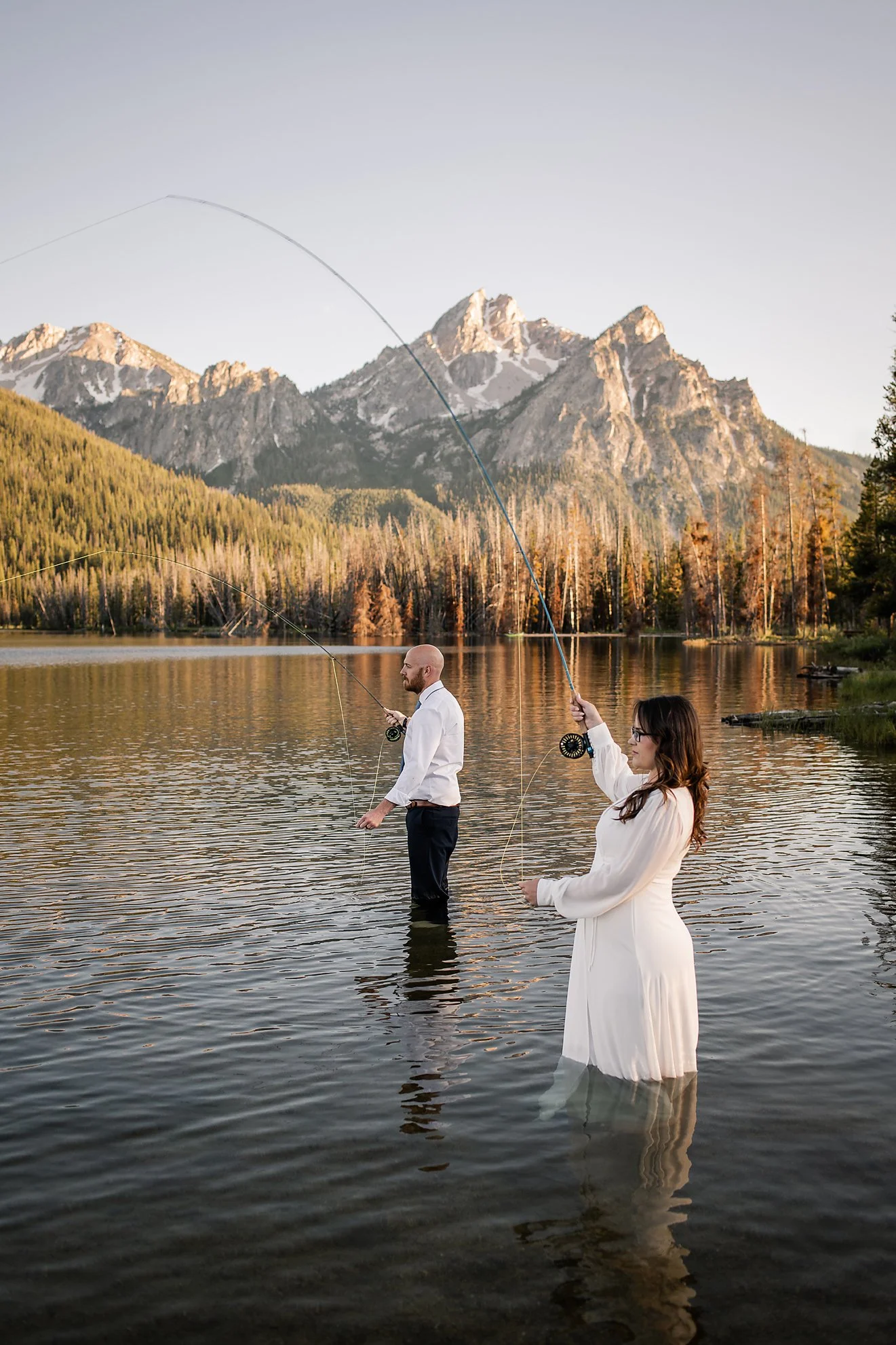 Bride and groom fly fishing in wedding attire at an alpine lake in the Sawtooth Mountains, during an adventurous Idaho elopement at sunset.