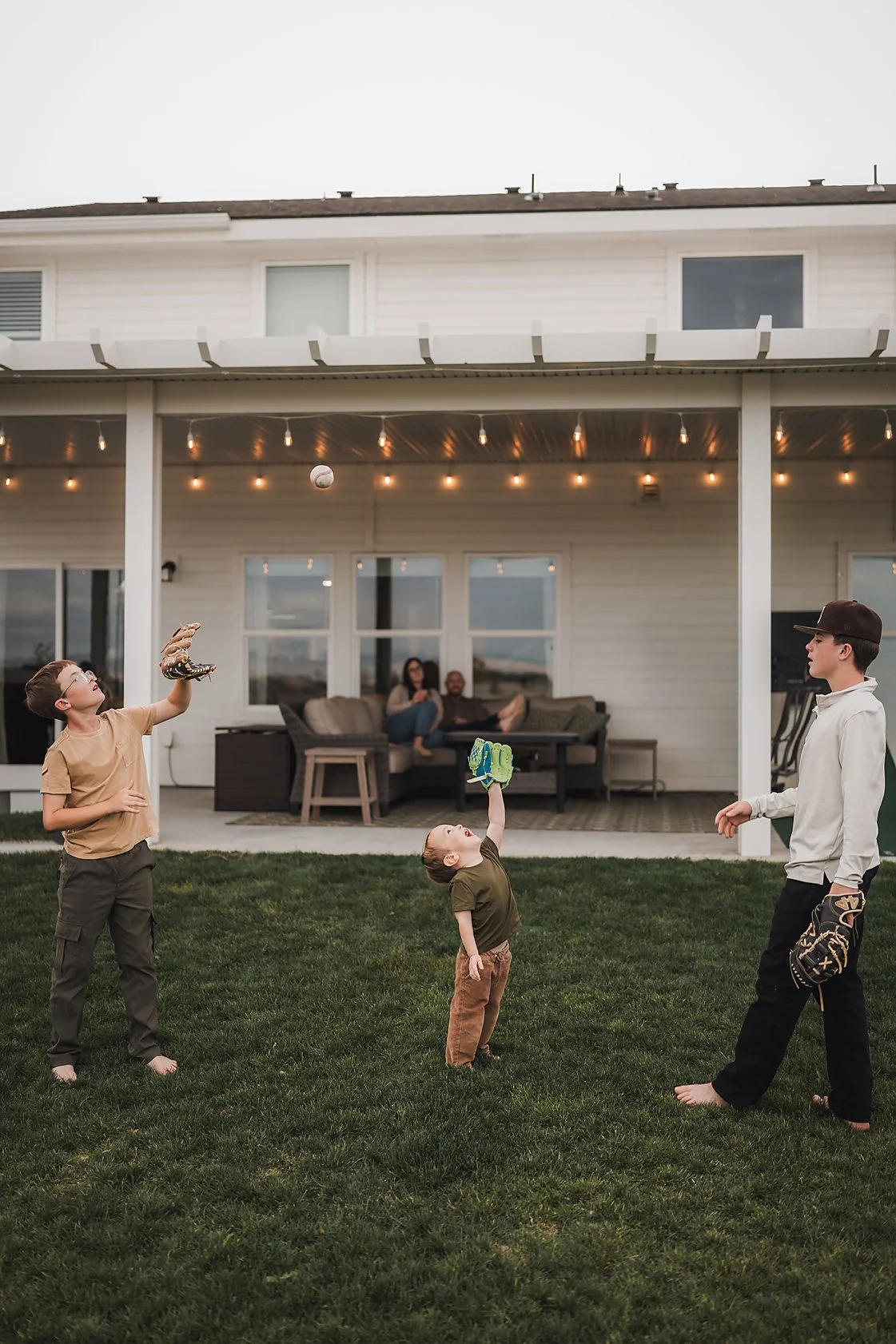 Children playing catch in the backyard while their parents relax on the patio during an in-home family photography session in Boise, Idaho.