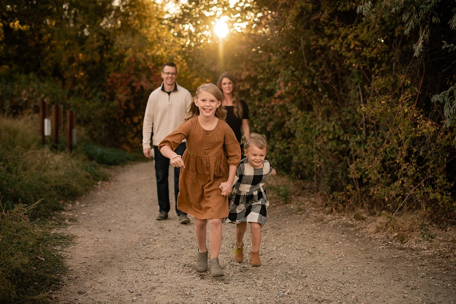 Two young sisters running down a wooded path during a Boise fall family session, parents walking behind them at sunset.
