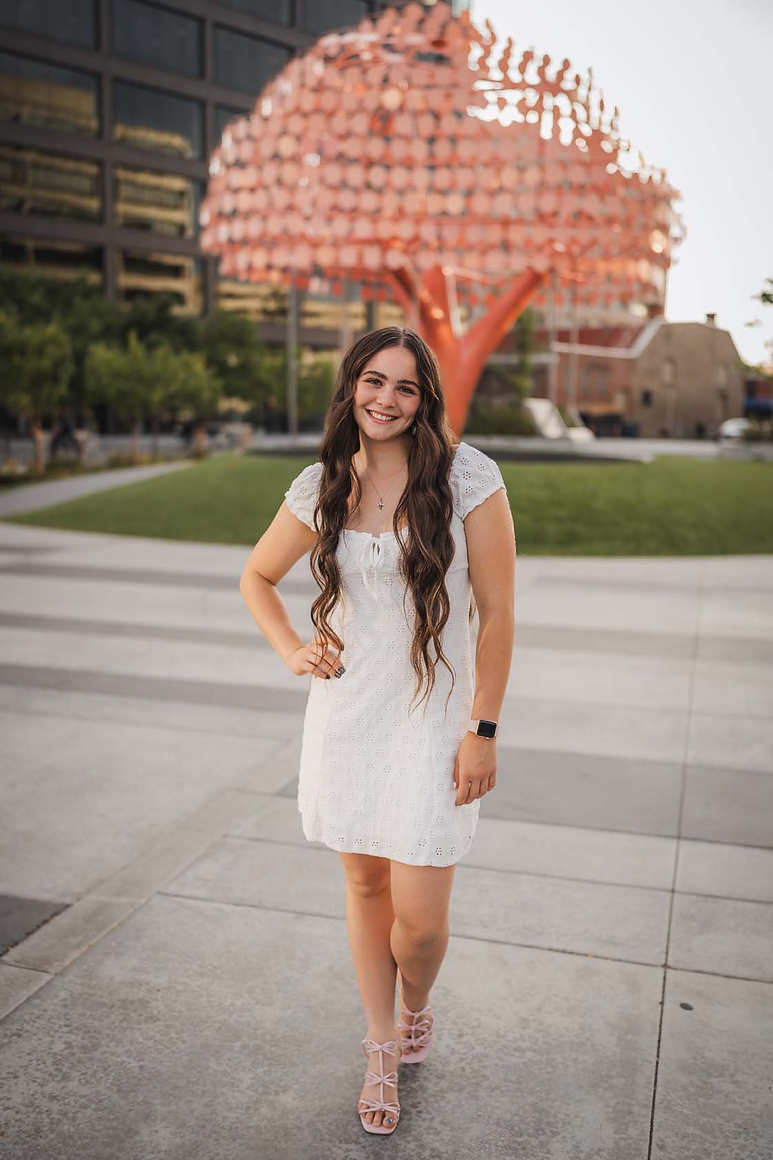 Eagle High School senior girl in a white dress walking through a downtown Boise Idaho park with the pink tree sculpture behind her