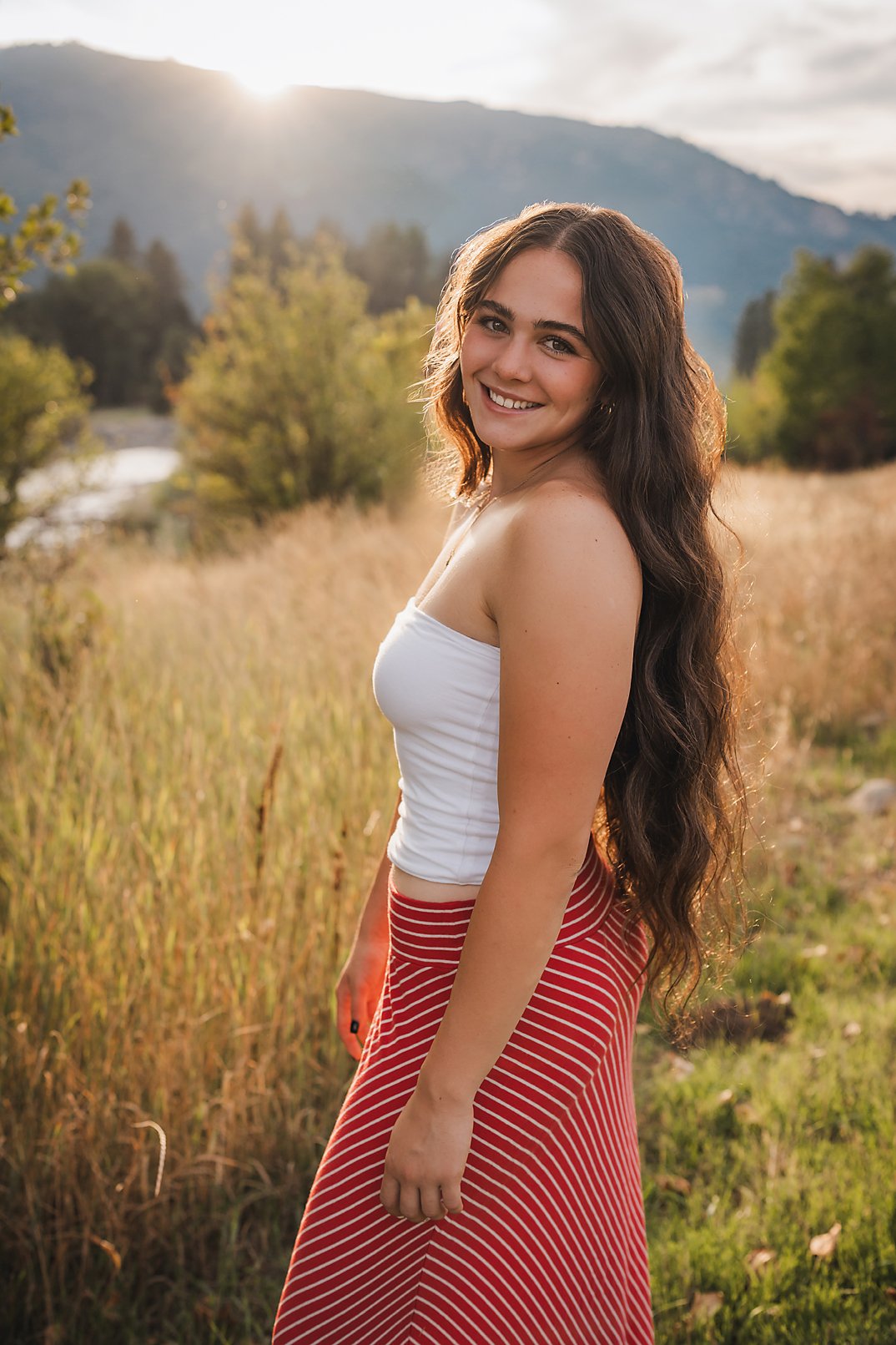 Eagle High School senior girl in a white top and red skirt standing in a golden field with mountains and sunset behind her in Garden Valley Idaho