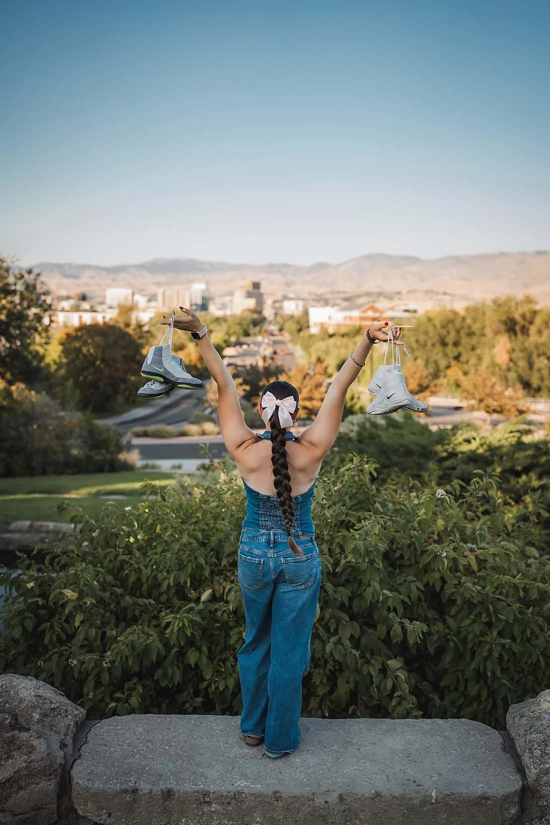 Eagle High School senior wrestler with a long braid and pink bow raising her wrestling shoes triumphantly overhead with a Boise Idaho city view behind her
