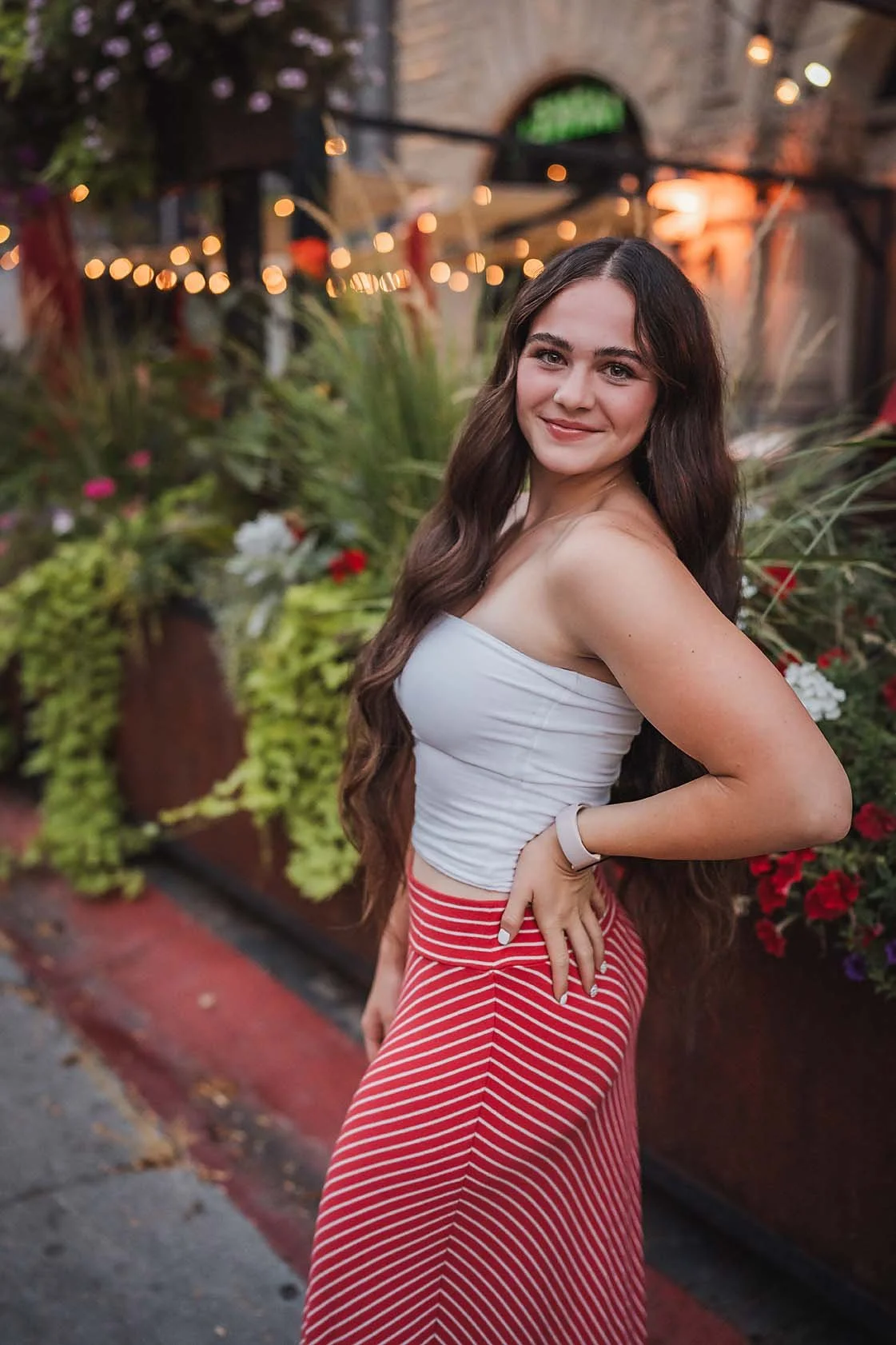 Eagle High School senior girl in a white top and red skirt smiling in front of flowers and cafe string lights in downtown Boise Idaho